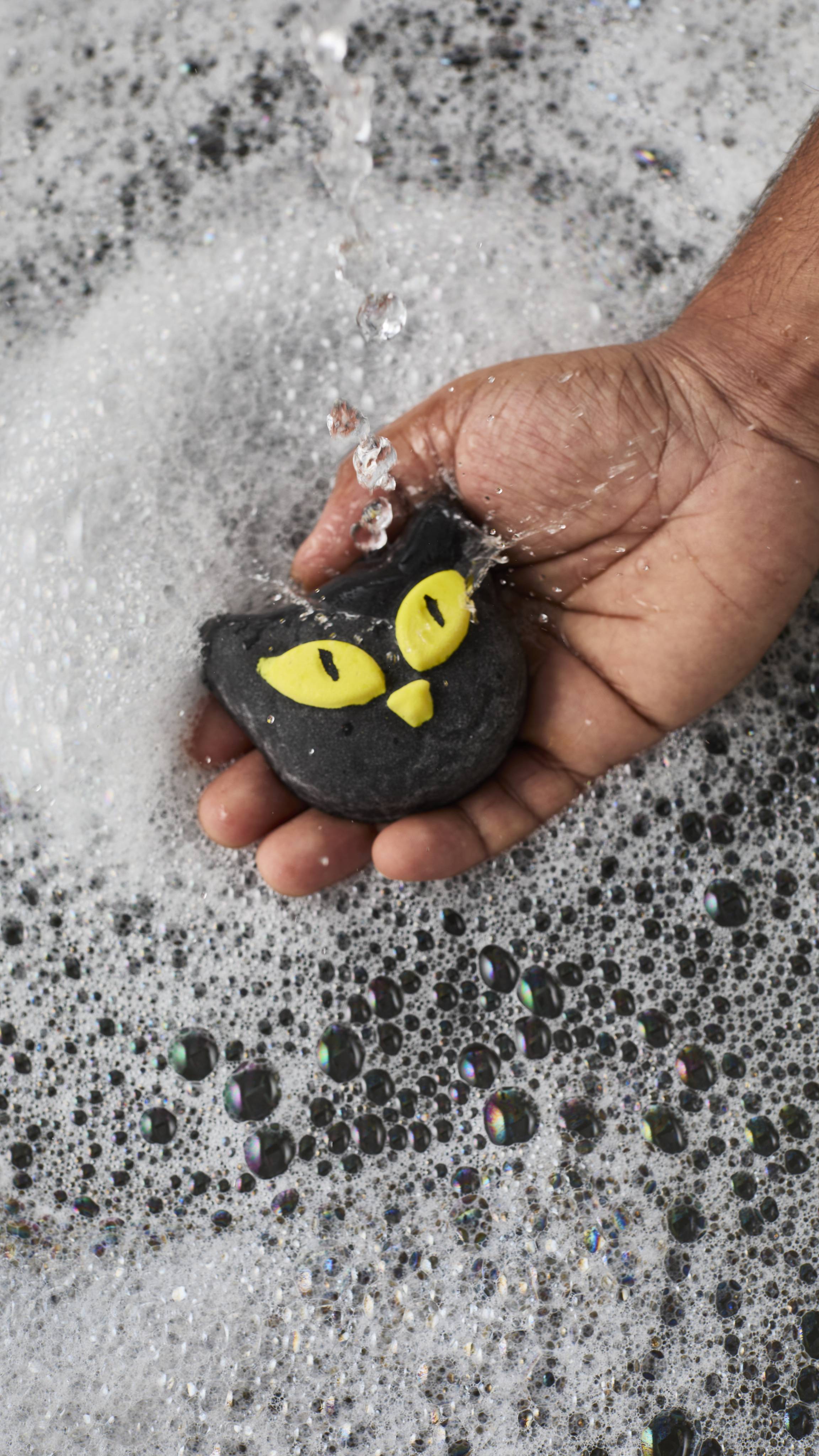 A person's hand holds a black cat-shaped bubble bar with bright yellow eyes under a stream of running tap water. The water splashes off the bar into a bath covered with a thick, frothy layer of white bubbles.