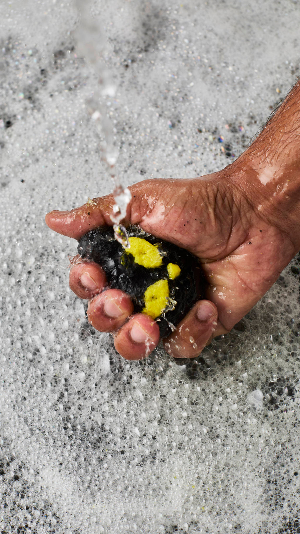A person's hand crumbles a black and yellow cat-shaped bubble bar under running tap water. The water splashes over the hand and into a bath filled with a dense, textured layer of white, frothy bubbles.