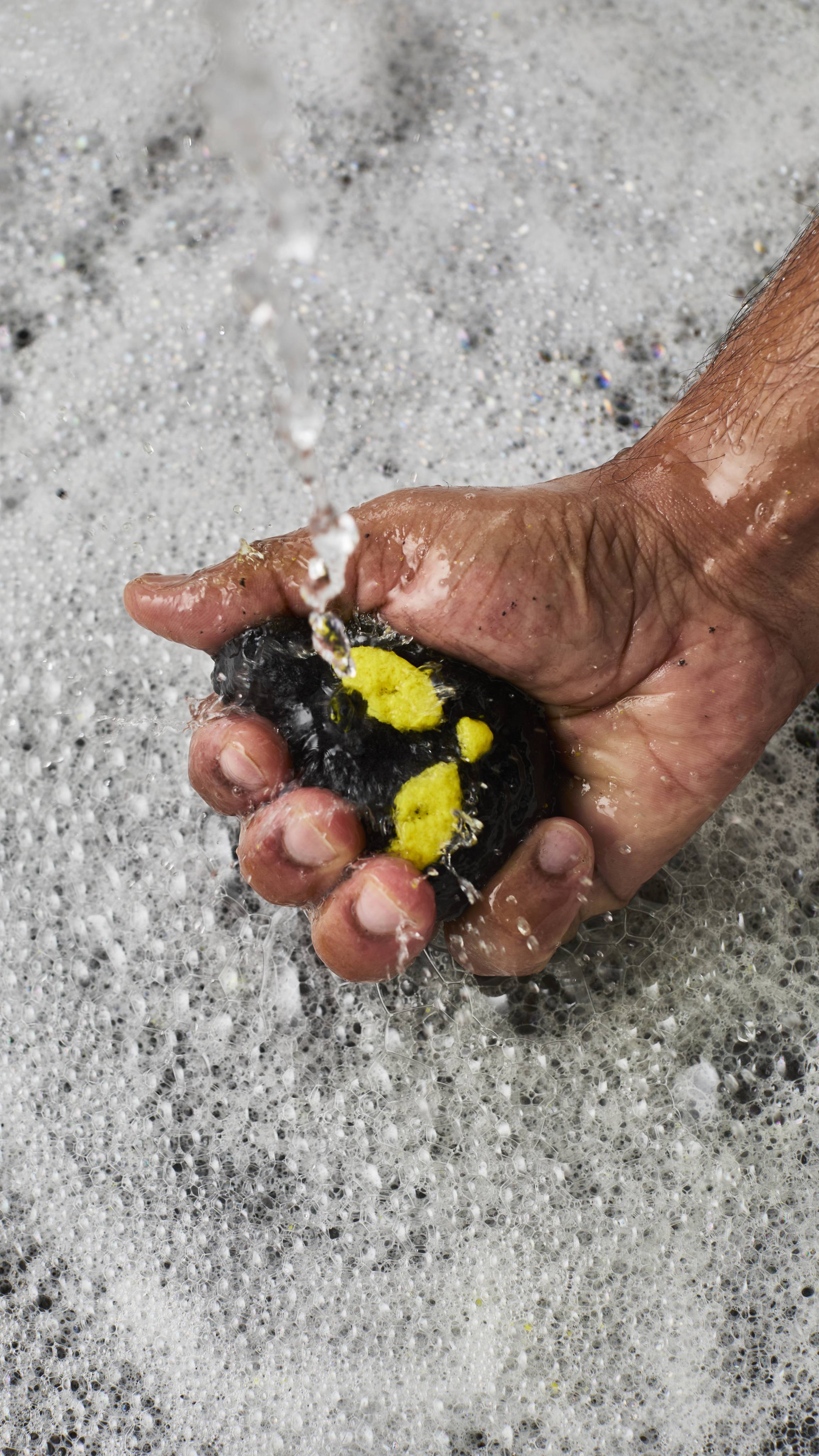 A person's hand crumbles a black and yellow cat-shaped bubble bar under running tap water. The water splashes over the hand and into a bath filled with a dense, textured layer of white, frothy bubbles.