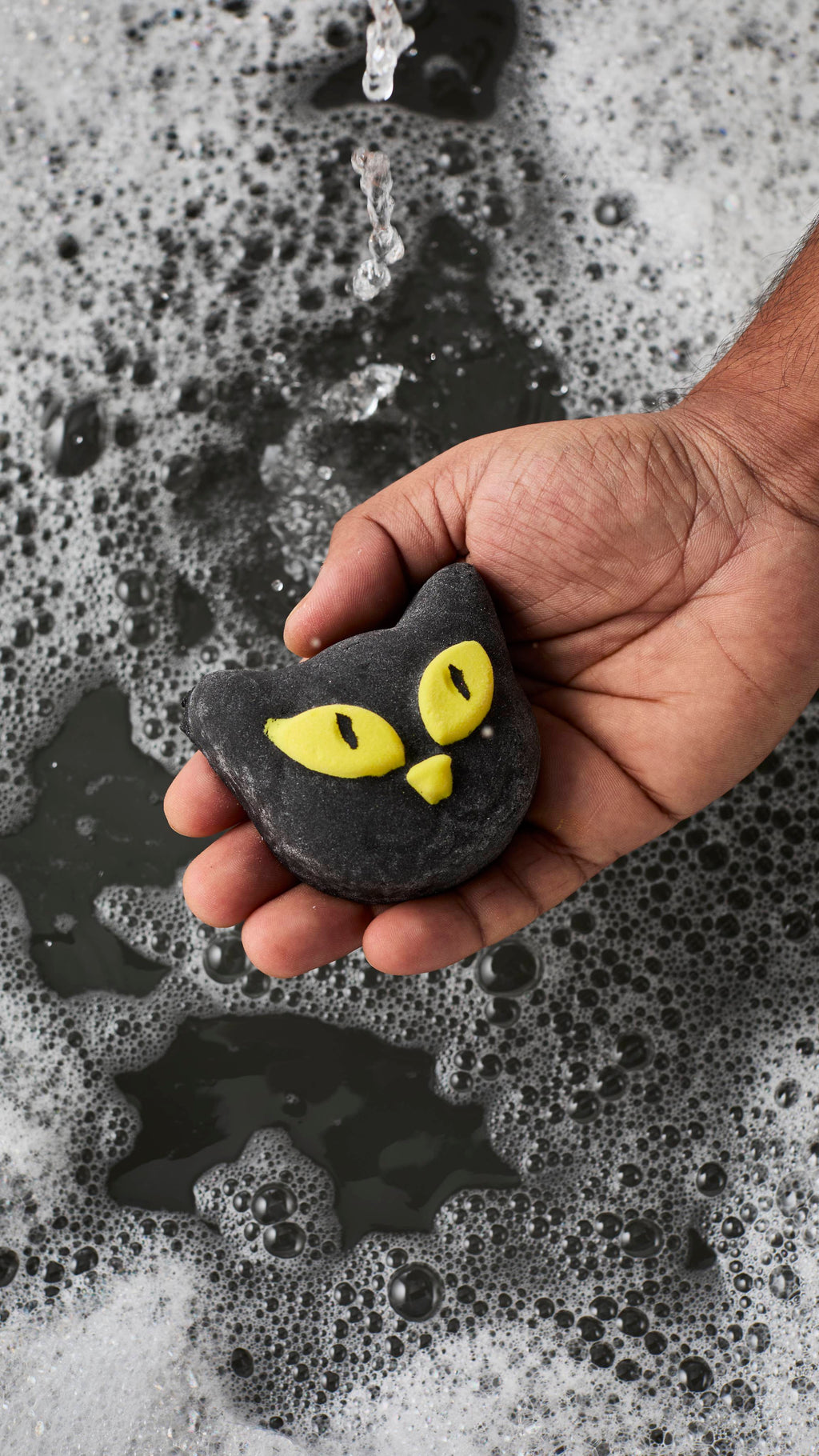 A person's hand holds a black cat-shaped bubble bar with bright yellow eyes and a nose over a bath. Running water splashes into the dark bath water below, which is covered in a dense layer of white, frothy bubbles.