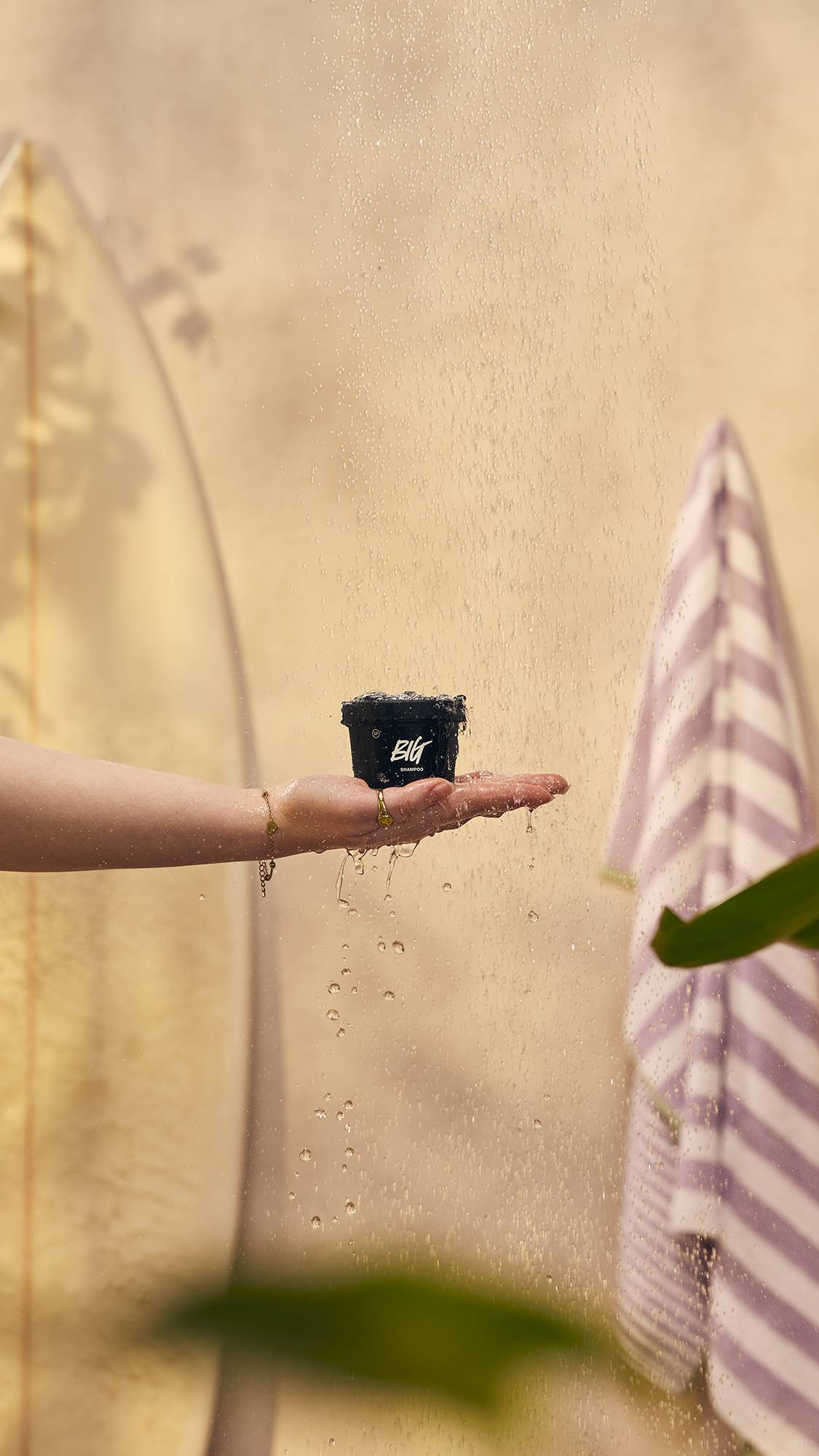 An outstretched hand holds a small black Lush pot labelled 'Big shampoo' under a spray of falling water. In the warm, neutral background, a surfboard rests on the left and a striped towel hangs on the right.