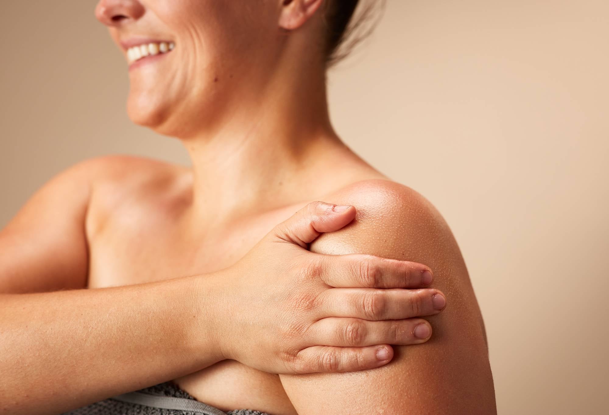 A smiling person wrapped in a towel gently rubs traces of a cream coloured body lotion, into their shoulder.