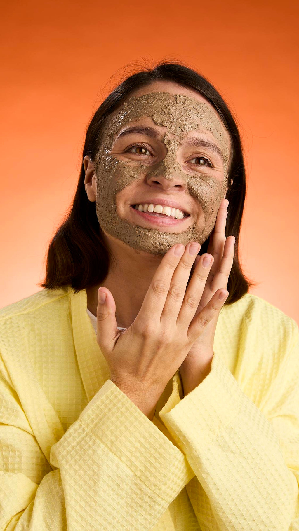 A smiling person with dark hair wears a yellow robe and touches their face, showcasing the full application and texture of the Black Tea Mochi face mask against an orange background.