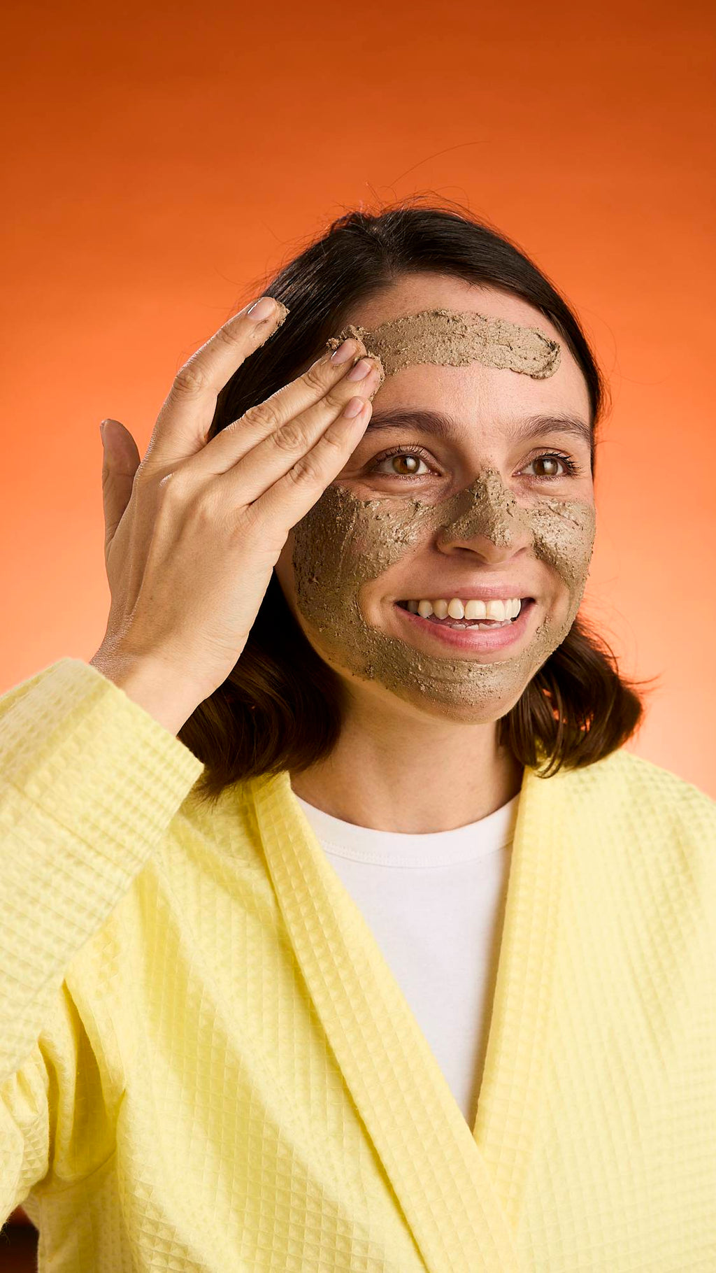 A smiling person wearing a yellow robe applies the thick, textured Black Tea Mochi face mask to their skin against a bright orange background.