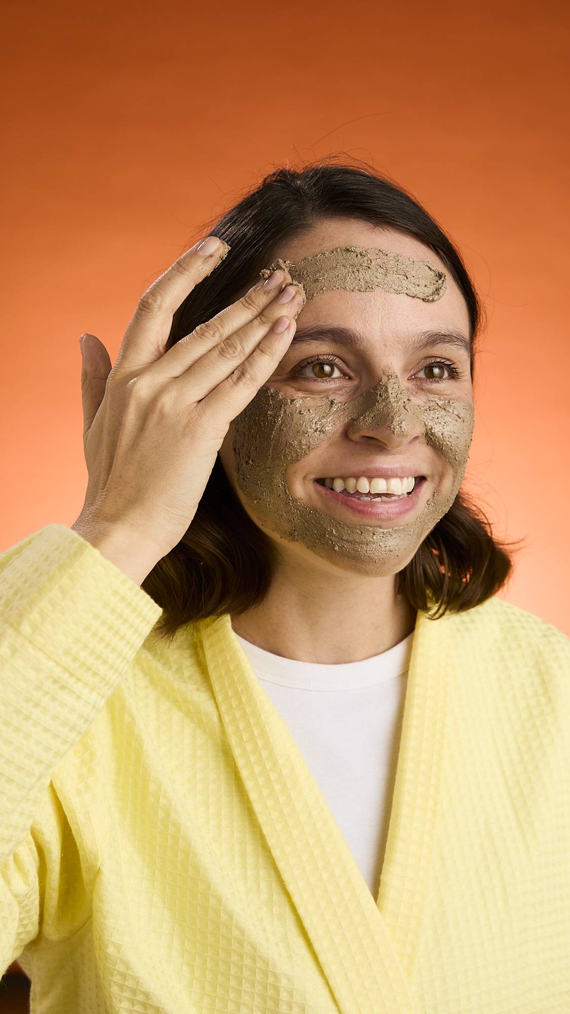 A smiling person wearing a yellow robe applies the thick, textured Black Tea Mochi face mask to their skin against a bright orange background.
