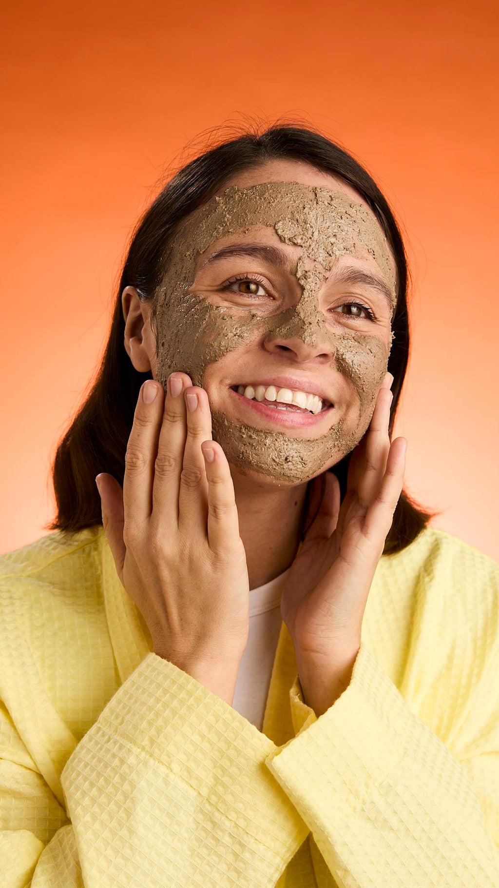 A smiling person with dark hair wears a yellow robe and touches their cheeks, showcasing the full application and texture of the Black Tea Mochi face mask against an orange background.