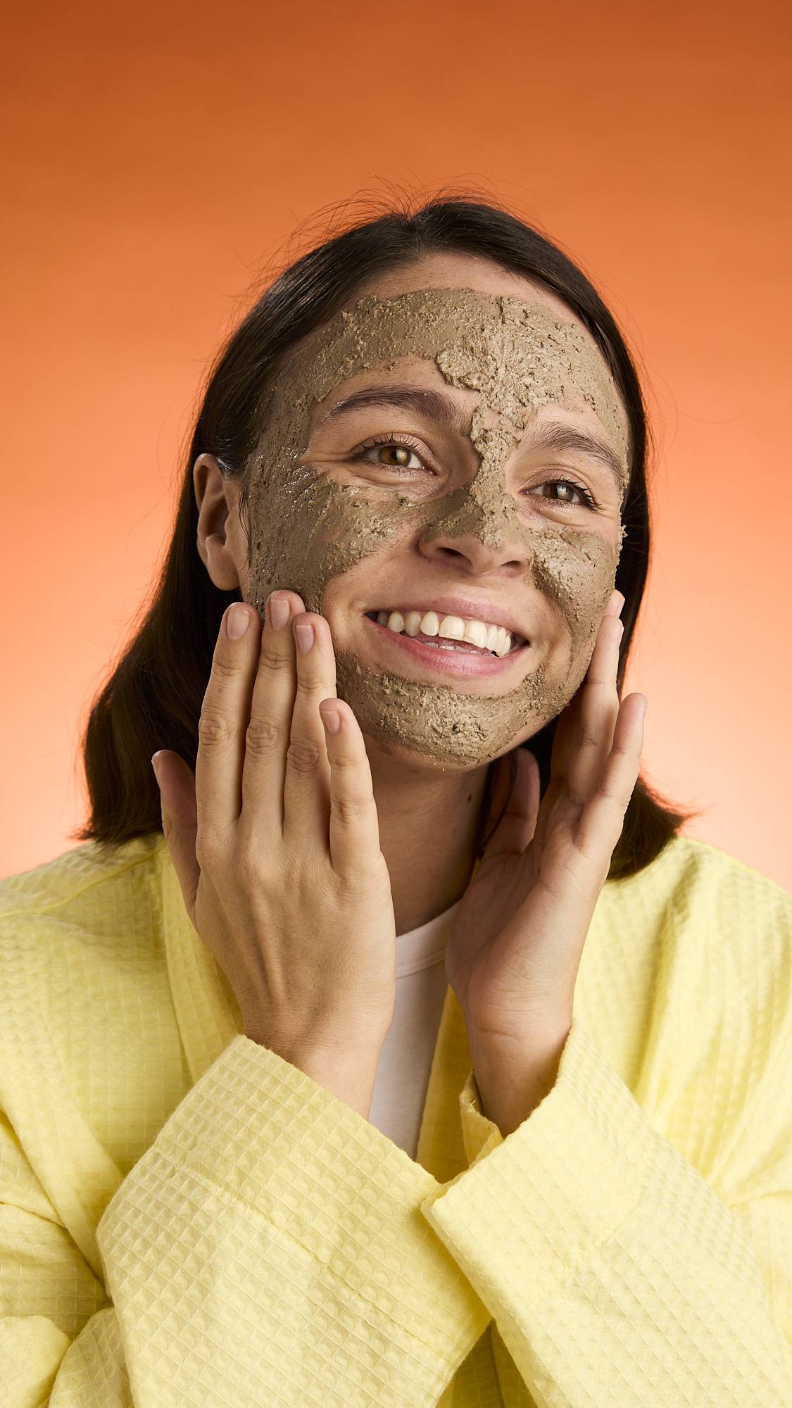 A smiling person with dark hair wears a yellow robe and touches their cheeks, showcasing the full application and texture of the Black Tea Mochi face mask against an orange background.