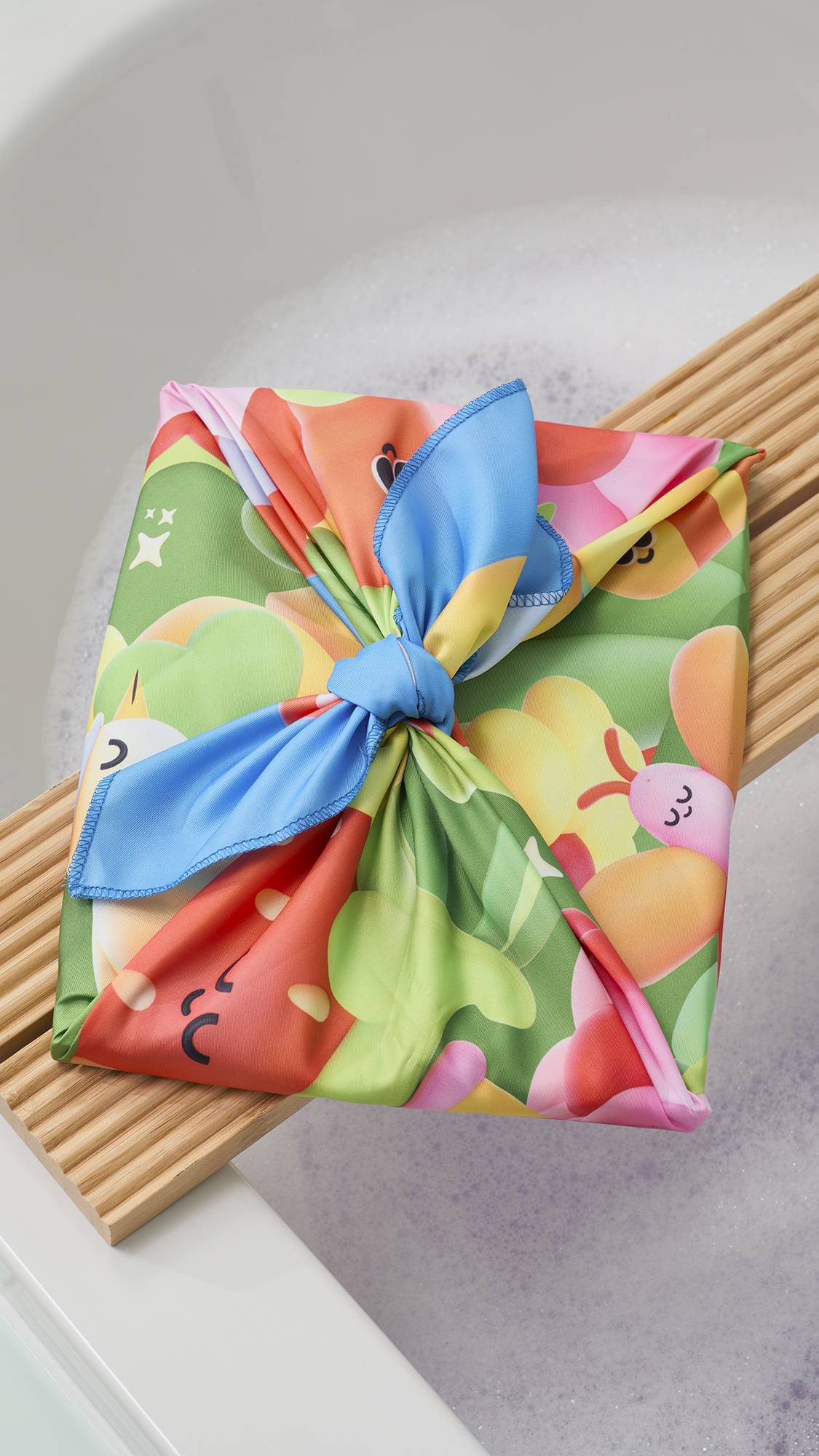 A vibrant "Blooming Lovely" fabric Knot Wrap, tied in a decorative bow, rests on a wooden bath trap over a soapy bathtub. The fabric features a colourful, 3D-style garden print with a large red strawberry and green foliage.