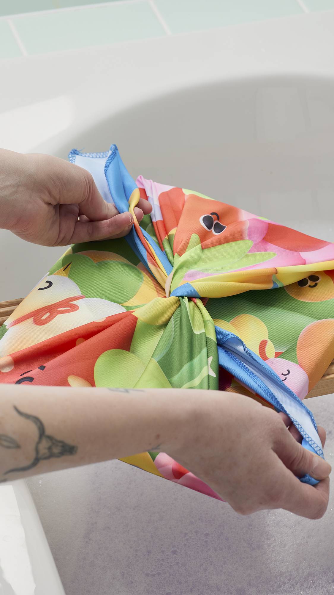 A model's hands are shown tying a "Blooming Lovely" Knot Wrap into a bow over a gift. The fabric features a colourful, 3D-style garden print. The gift rests on a wooden bath tray over a soapy bathtub in a bright bathroom.