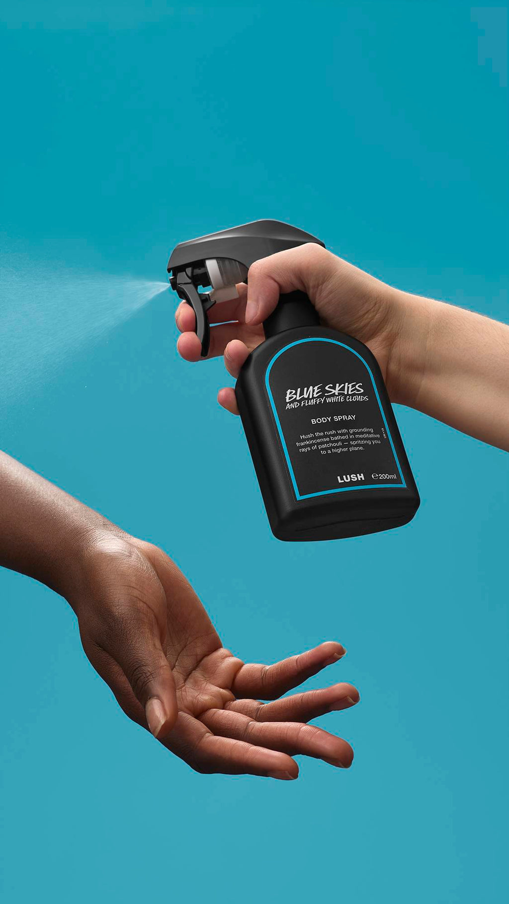 The image shows a light-toned hand spraying a bottle of LUSH 'Blue Skies and Fluffy White Clouds' body spray. A warm-toned hand is cupped below, catching the fine mist against a bright, sky-blue background.