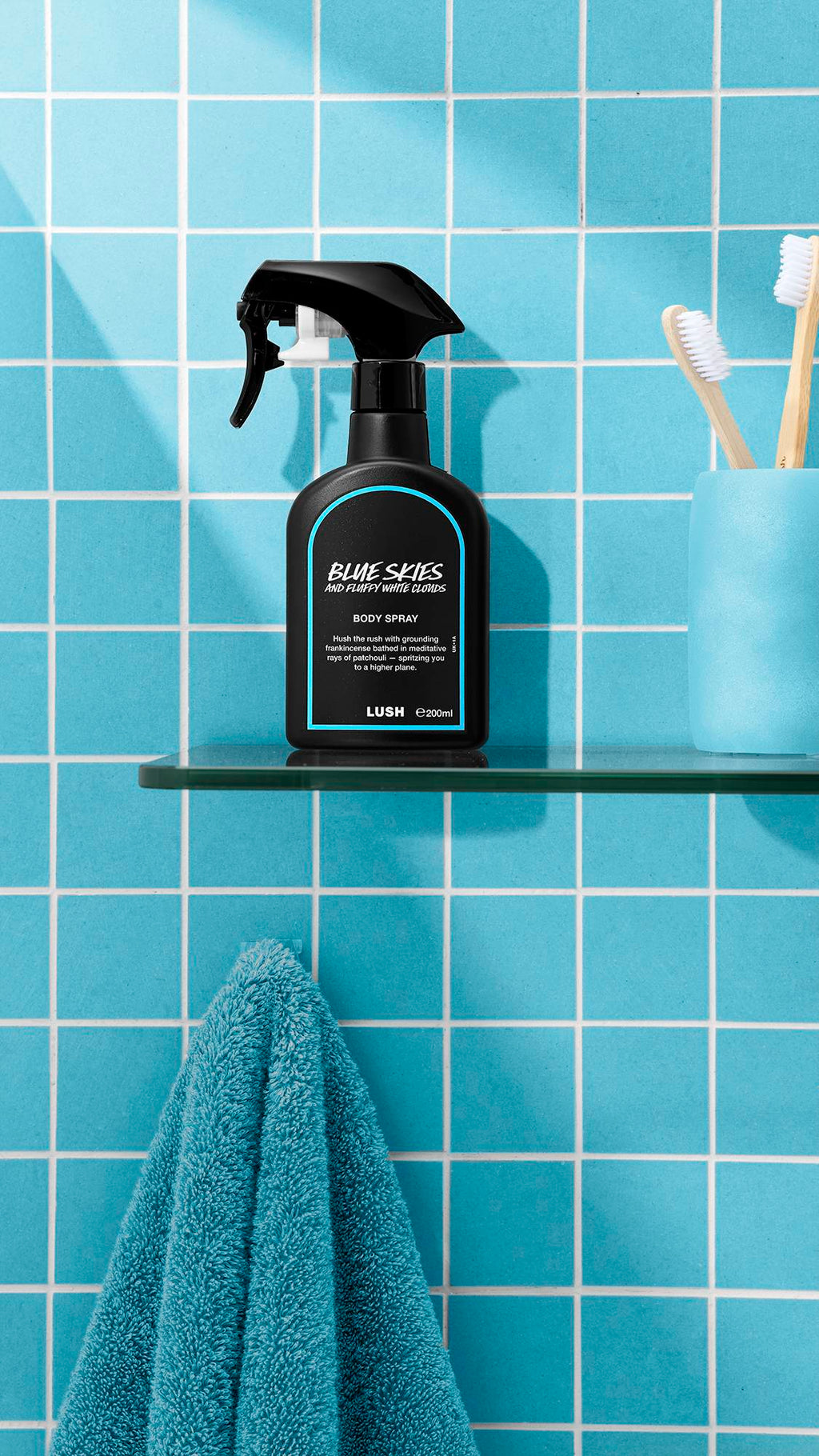 The image shows a bottle of LUSH 'Blue Skies and Fluffy White Clouds' body spray on a glass shelf. A teal towel hangs below, with a blue cup and brushes nearby, against a bright, tiled, sky-blue bathroom wall.