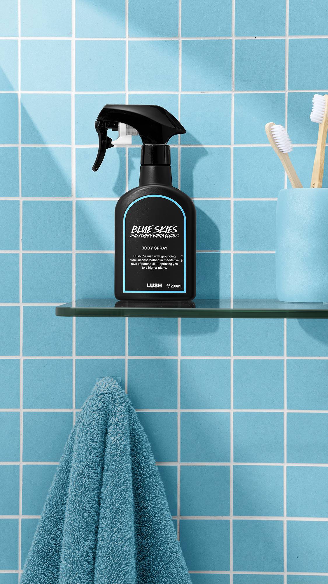 The image shows a bottle of LUSH 'Blue Skies and Fluffy White Clouds' body spray on a glass shelf. A teal towel hangs below, with a blue cup and brushes nearby, against a bright, tiled, sky-blue bathroom wall.