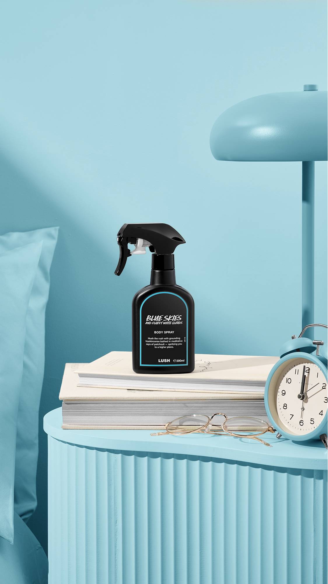 The image shows a recycled black plastic LUSH spray bottle of "Blue Skies and Fluffy White Clouds" sitting on a pile of books on a light blue bedside table. A matching lamp, alarm clock, and glasses sit nearby, next to a blue bed and wall.