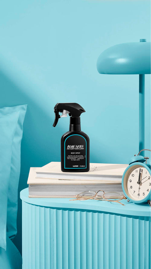The image shows a recycled black plastic LUSH spray bottle of "Blue Skies and Fluffy White Clouds" sitting on a pile of books on a light blue bedside table. A matching lamp, alarm clock, and glasses sit nearby, next to a blue bed and wall.