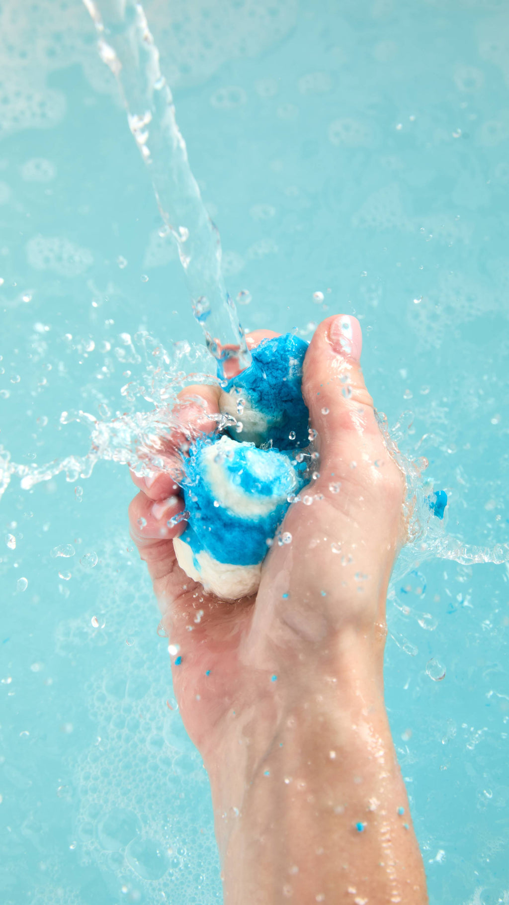 The image shows a light-toned hand vigorously squeezing and crumbling a blue and white 'Mini Blue Skies' bubble bar under a running tap. A strong stream creates vibrant blue bath water with abundant foam and bubbles.