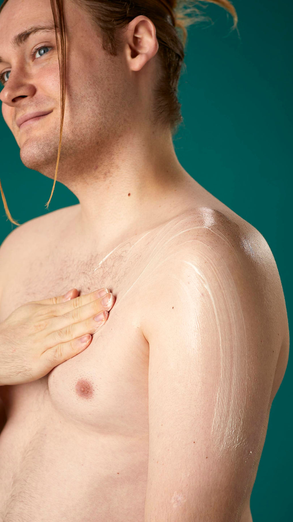 The image shows a topless model on a teal background. They are gently soothing the Braziliant body butter in circular motions over their chest and upper arm.