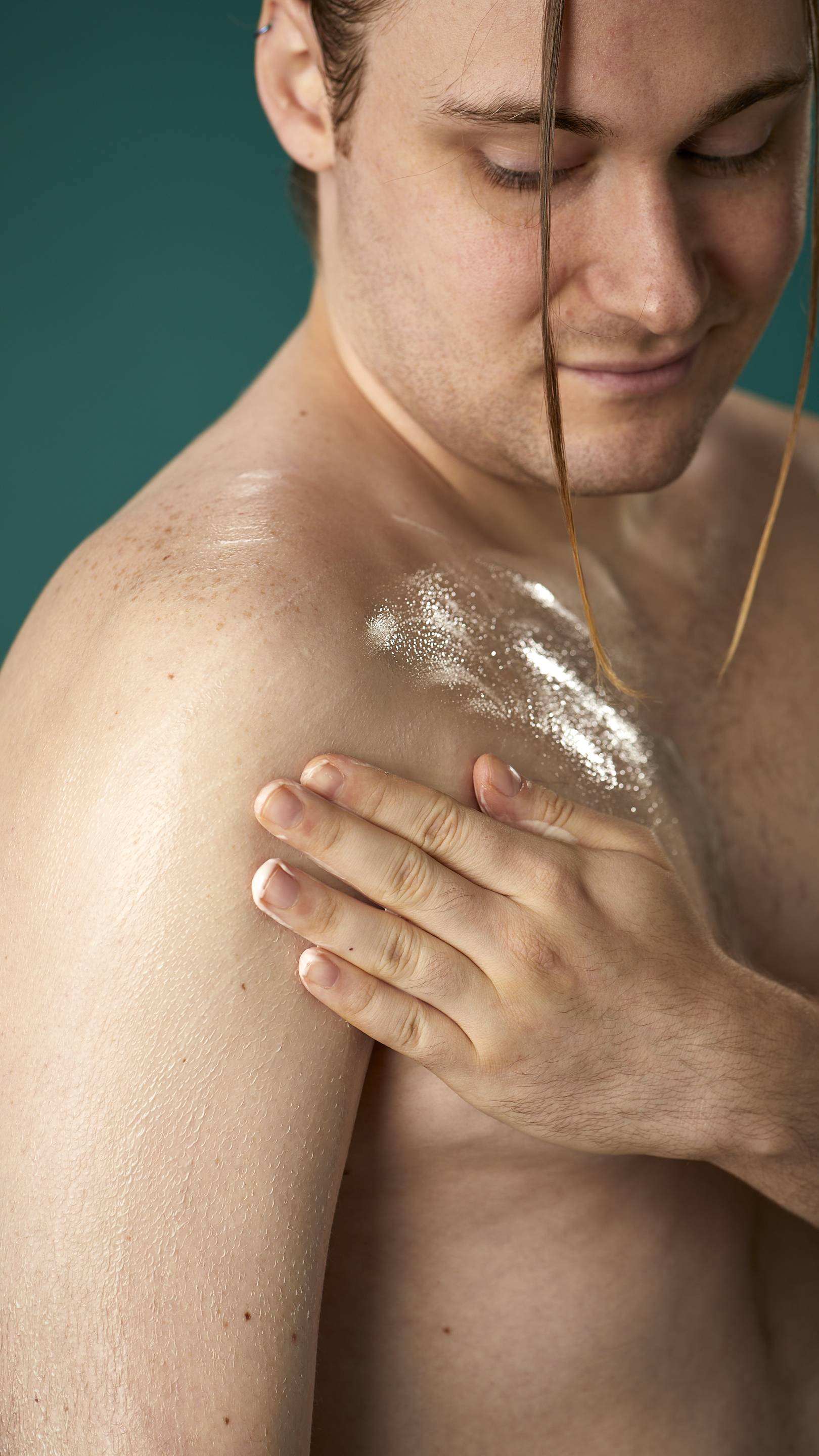 The image shows a topless model on a teal background. They are gently soothing the Braziliant body butter in circular motions over their chest and upper arm.