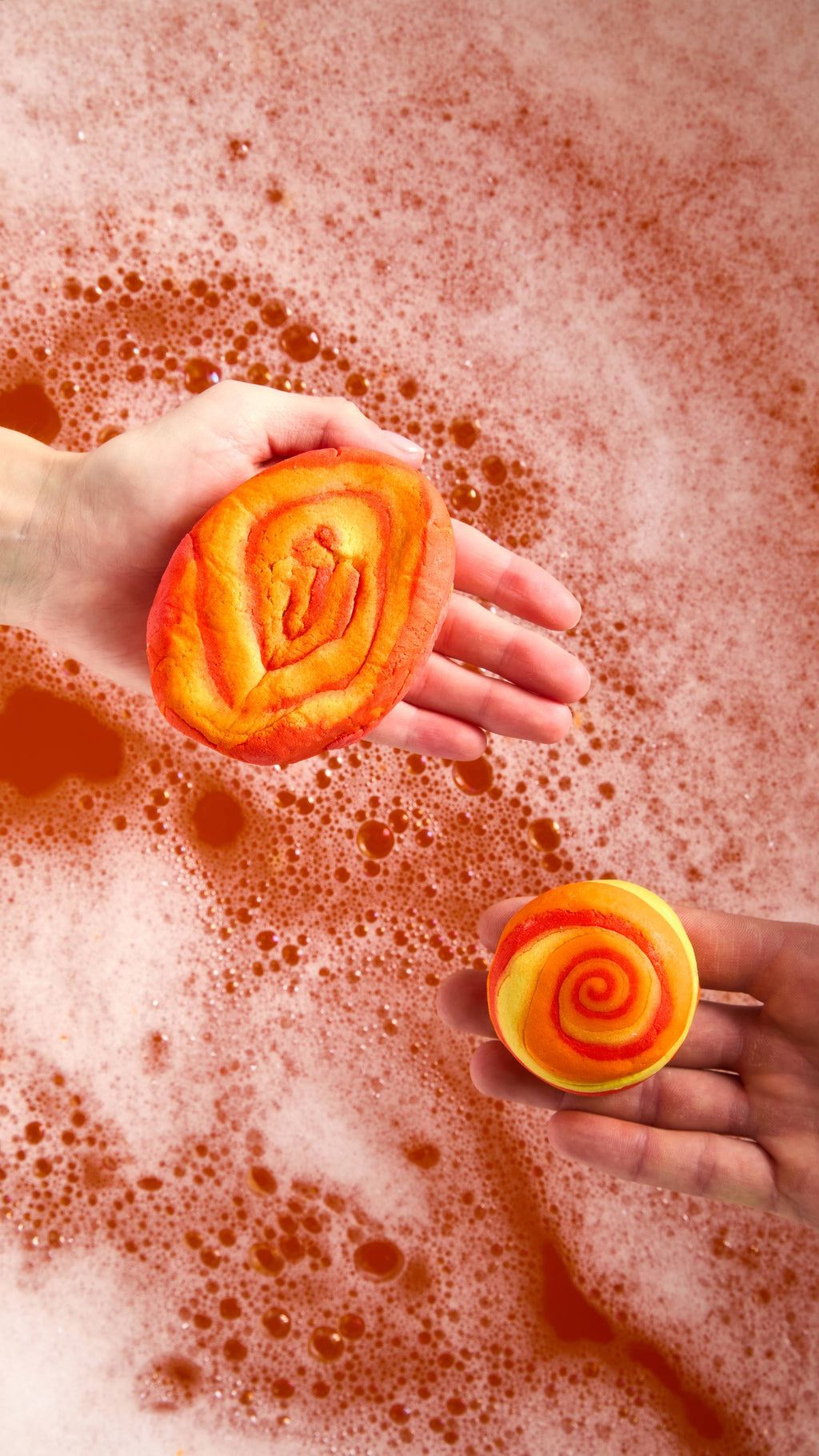 The image shows a close-up of two hands holding two different sizes of vibrant Brightside bubble bars above the firey orange, bubbly water below.