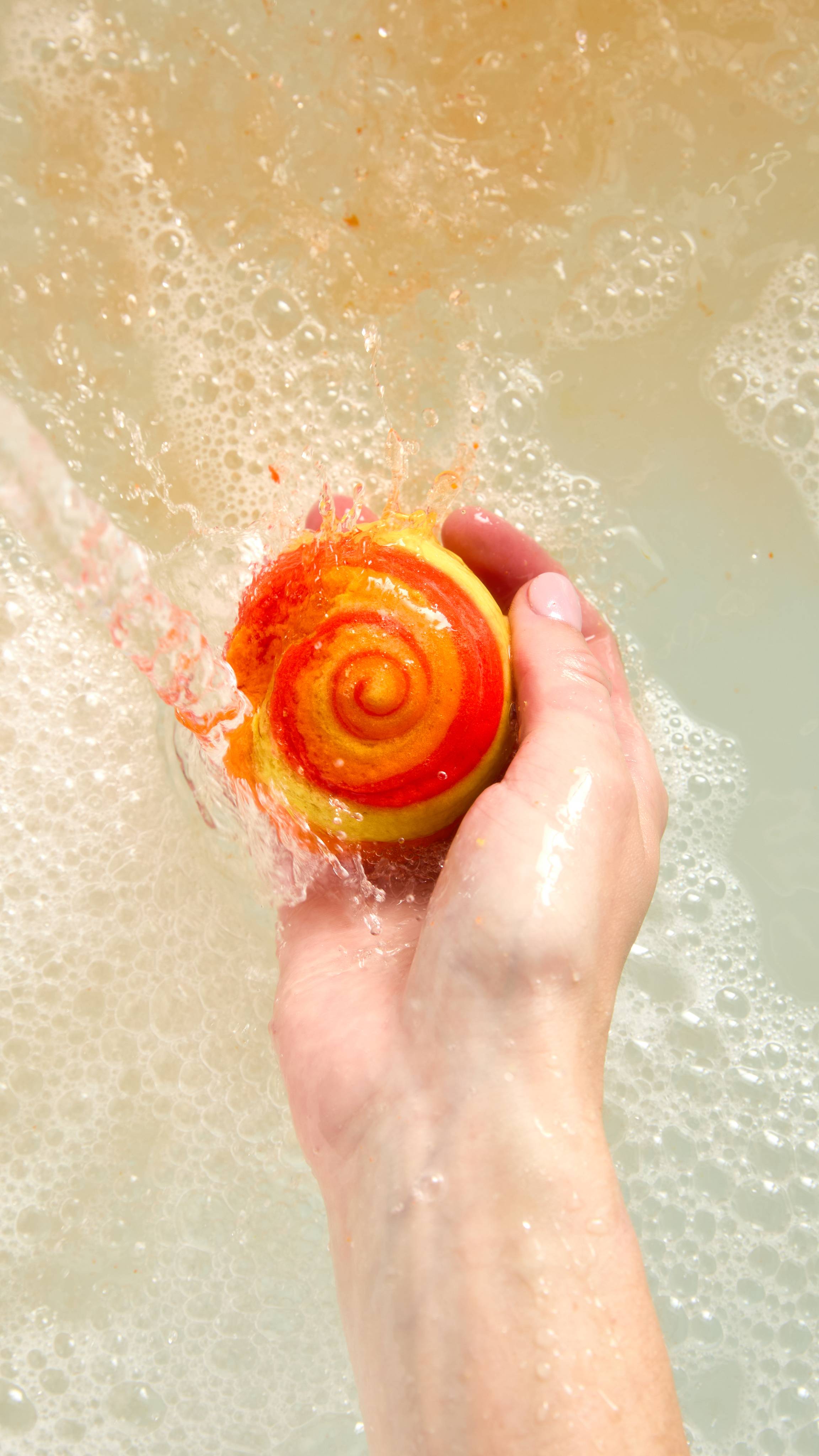 The image is an aerial view of the model's hand reaching in from below and holding the vibrant, orange Brightside mini bubble bar in the hand under running water into the bath water below. Underneath, the water has a layer of thick bubbles.