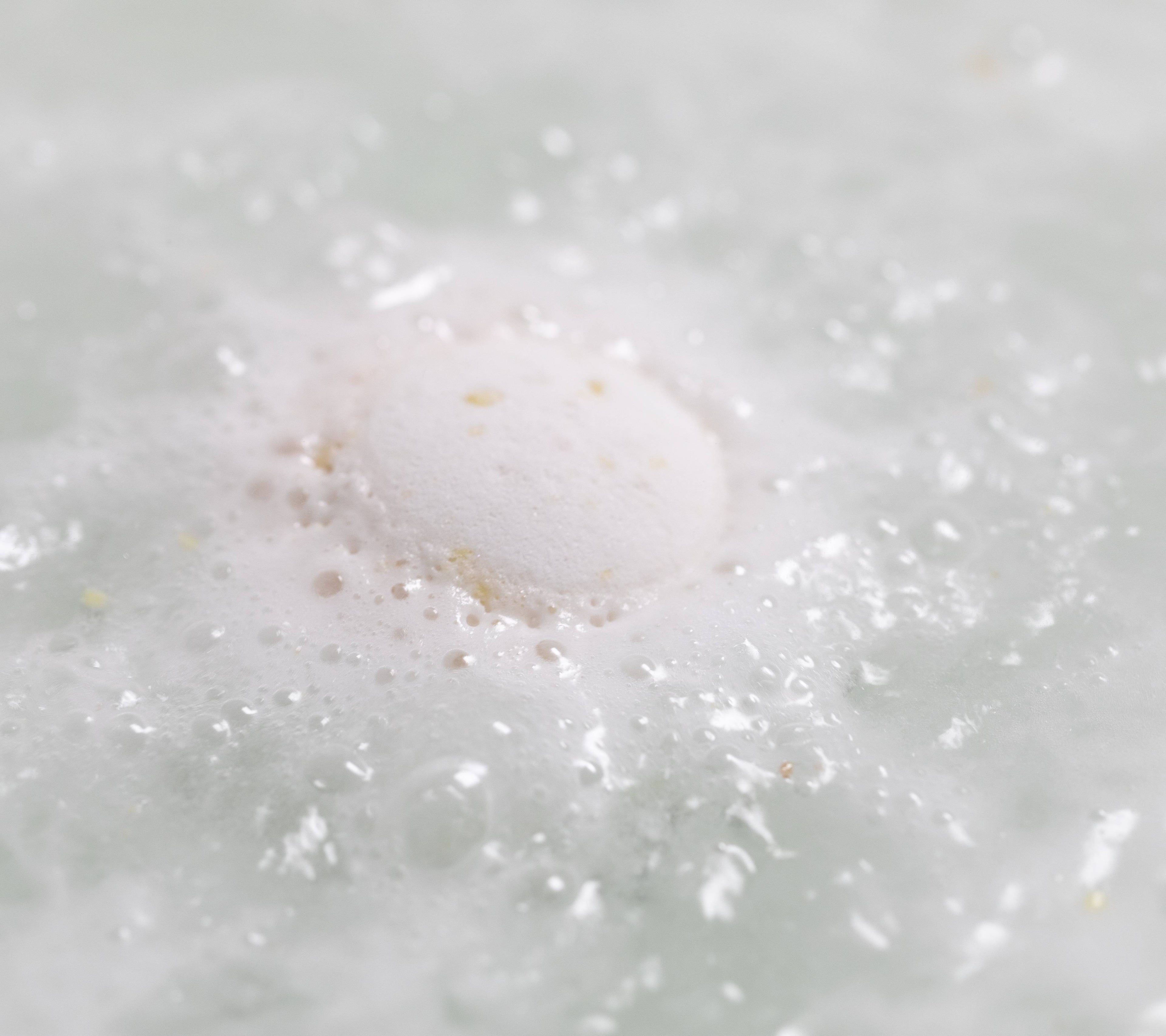 The image shows a close-up of the Butterball bath bomb. The white bath bomb dissolves in the water leaving behind delicate white foam and tiny chunks of solid cocoa butter.