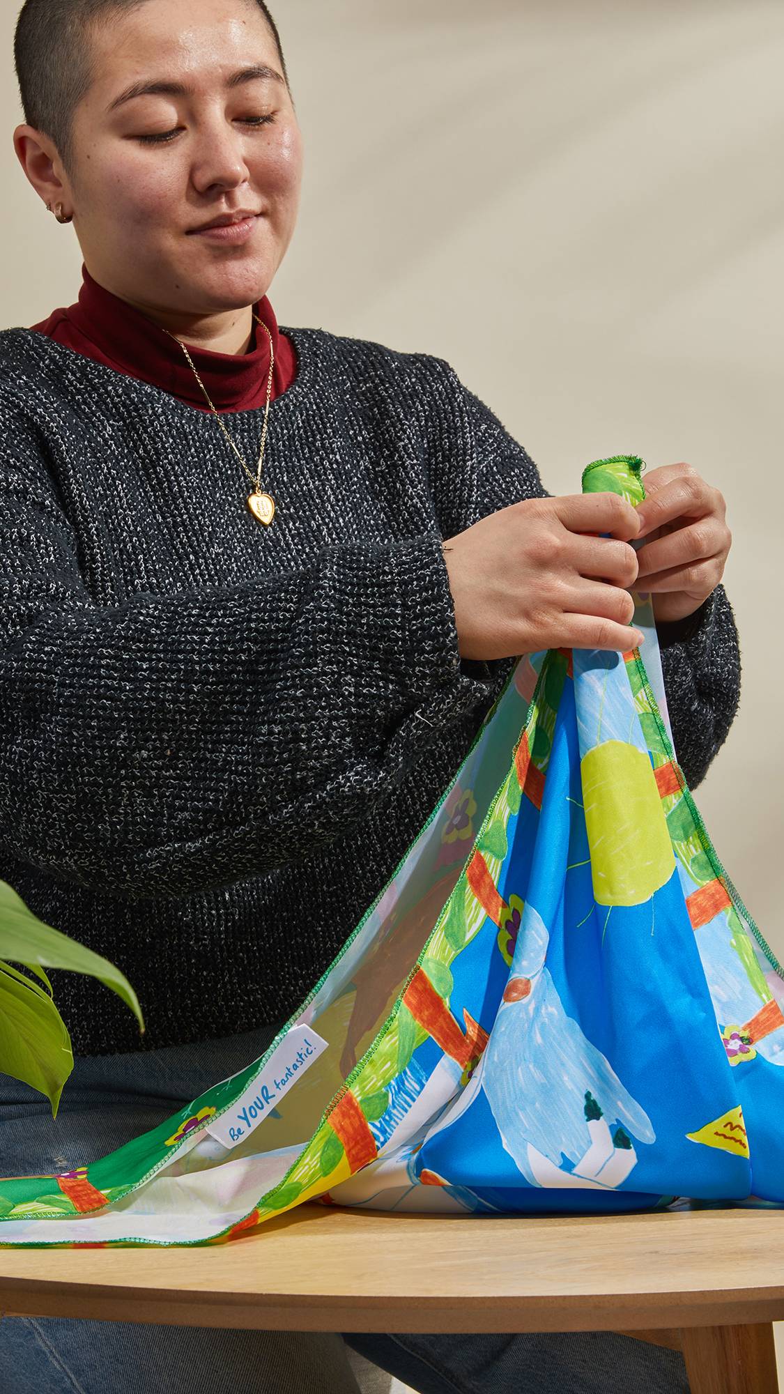 The image shows the model as they sit at a table and neatly tie the By The Seaside knot wrap around a gift by lifting the corners and bringing them together.