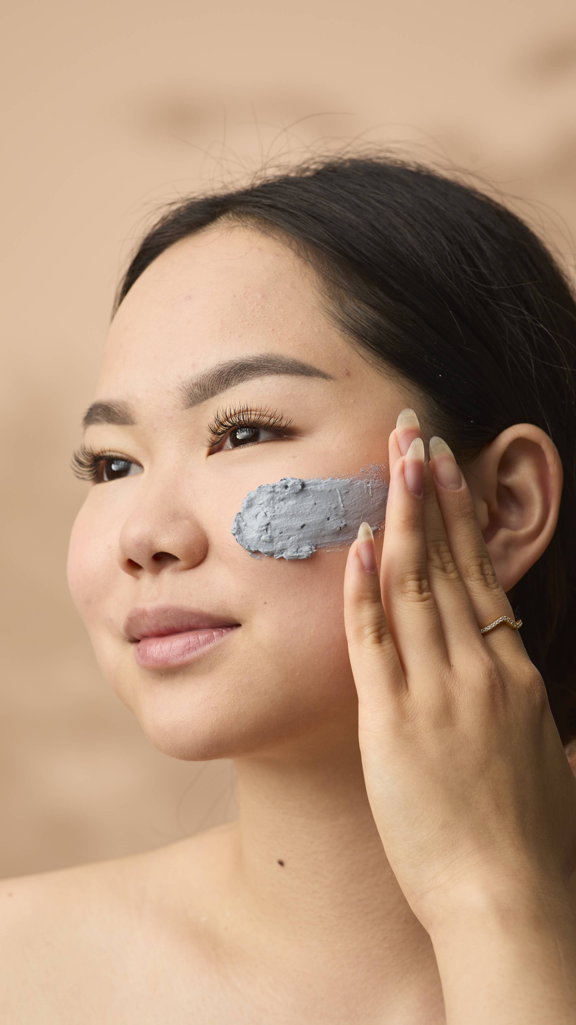 A close-up portrait of a woman applying Catastrophe Cosmetic face mask to her cheek. She uses her fingers to spread the textured cream across her skin. She has dark hair styled back, long natural fingernails, and is wearing a thin gold ring.