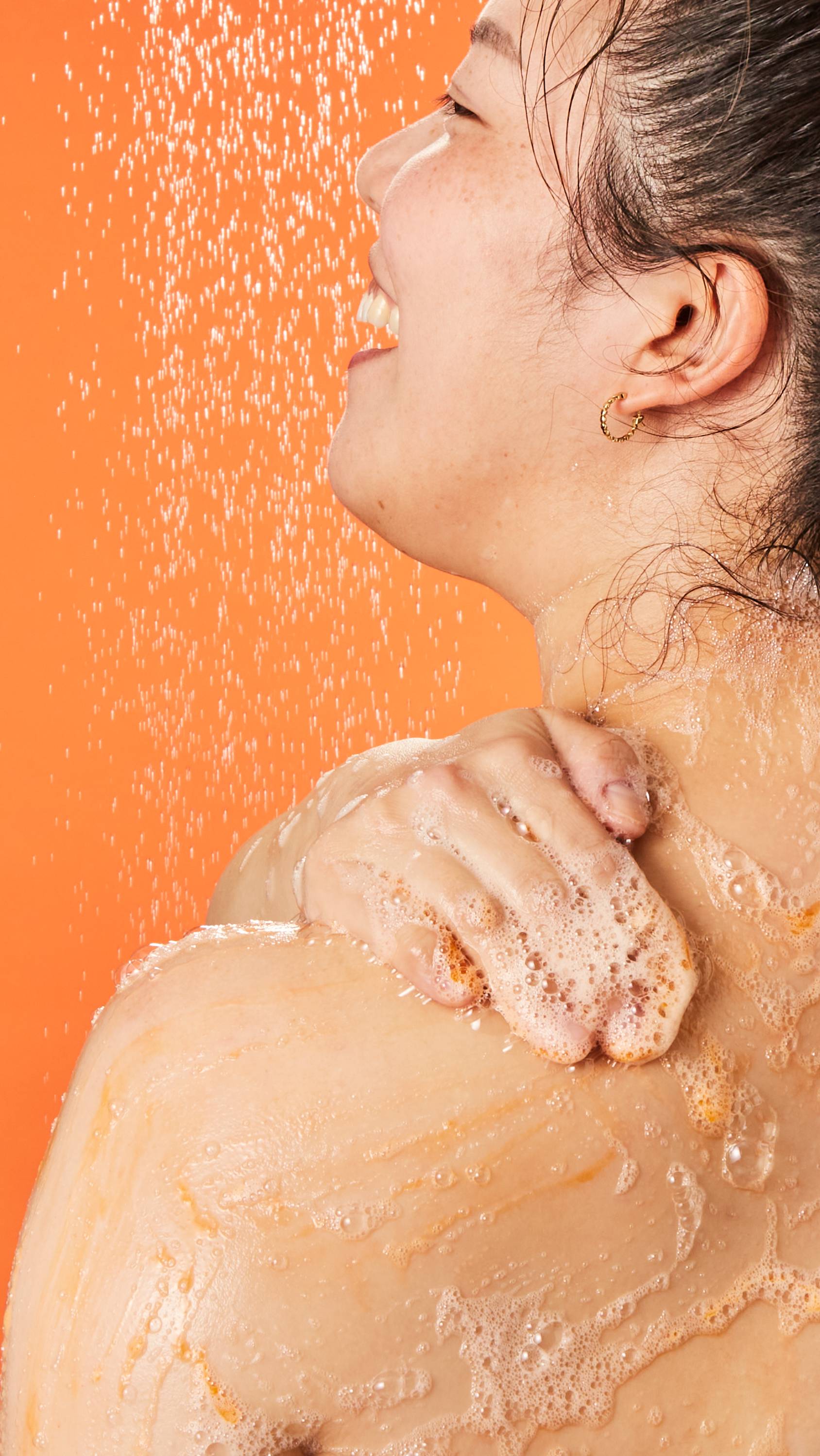 Image shows a close-up of the model's back as they face away. They are lathering up the shower gel on their shoulder.