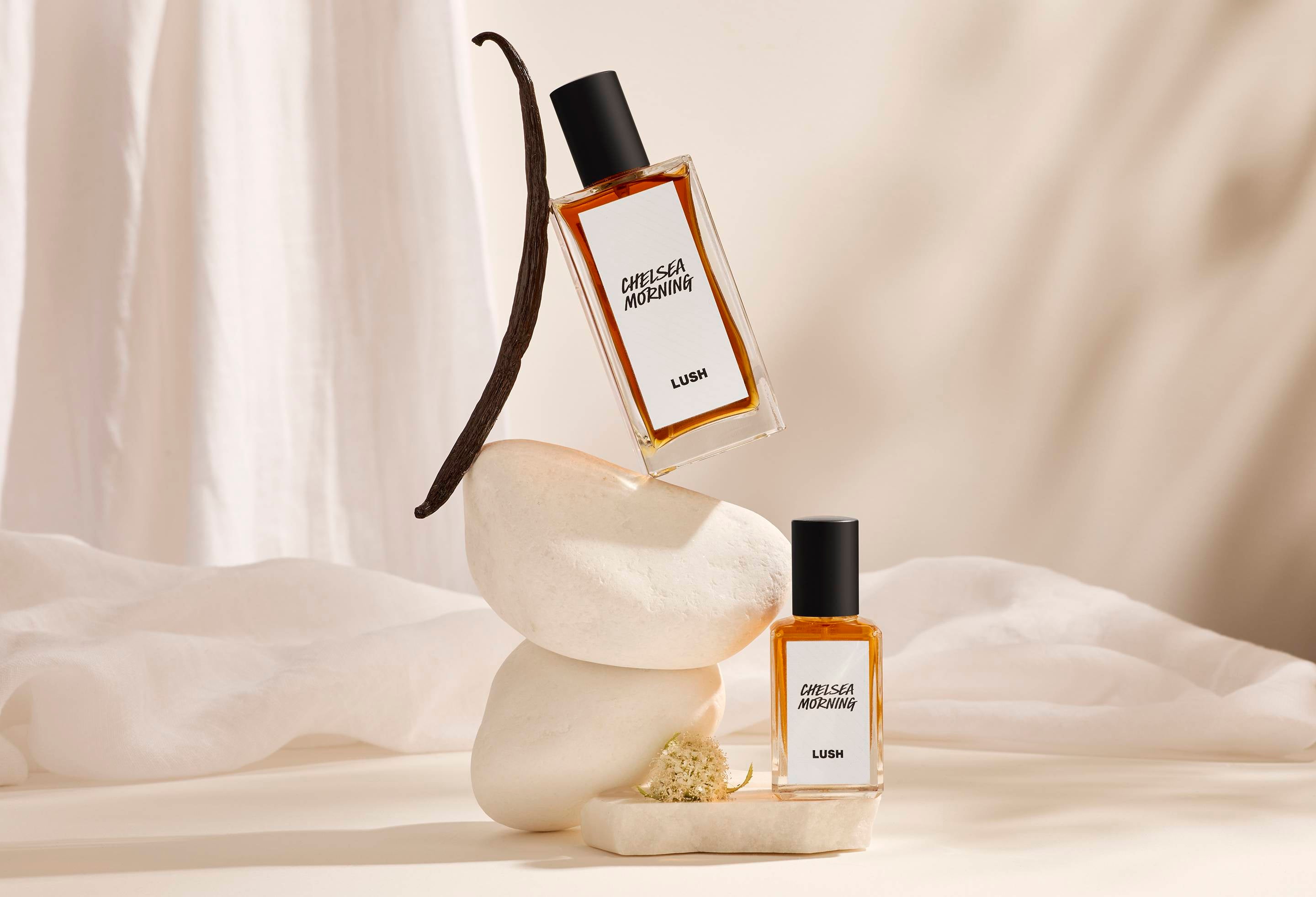 The image shows a background of layered, cream, satin fabric. In the centre is the focus of precisely balanced, white stacked stones. There is a dried vanilla pot standing on top and the two sizes of perfume leaning against the stones.