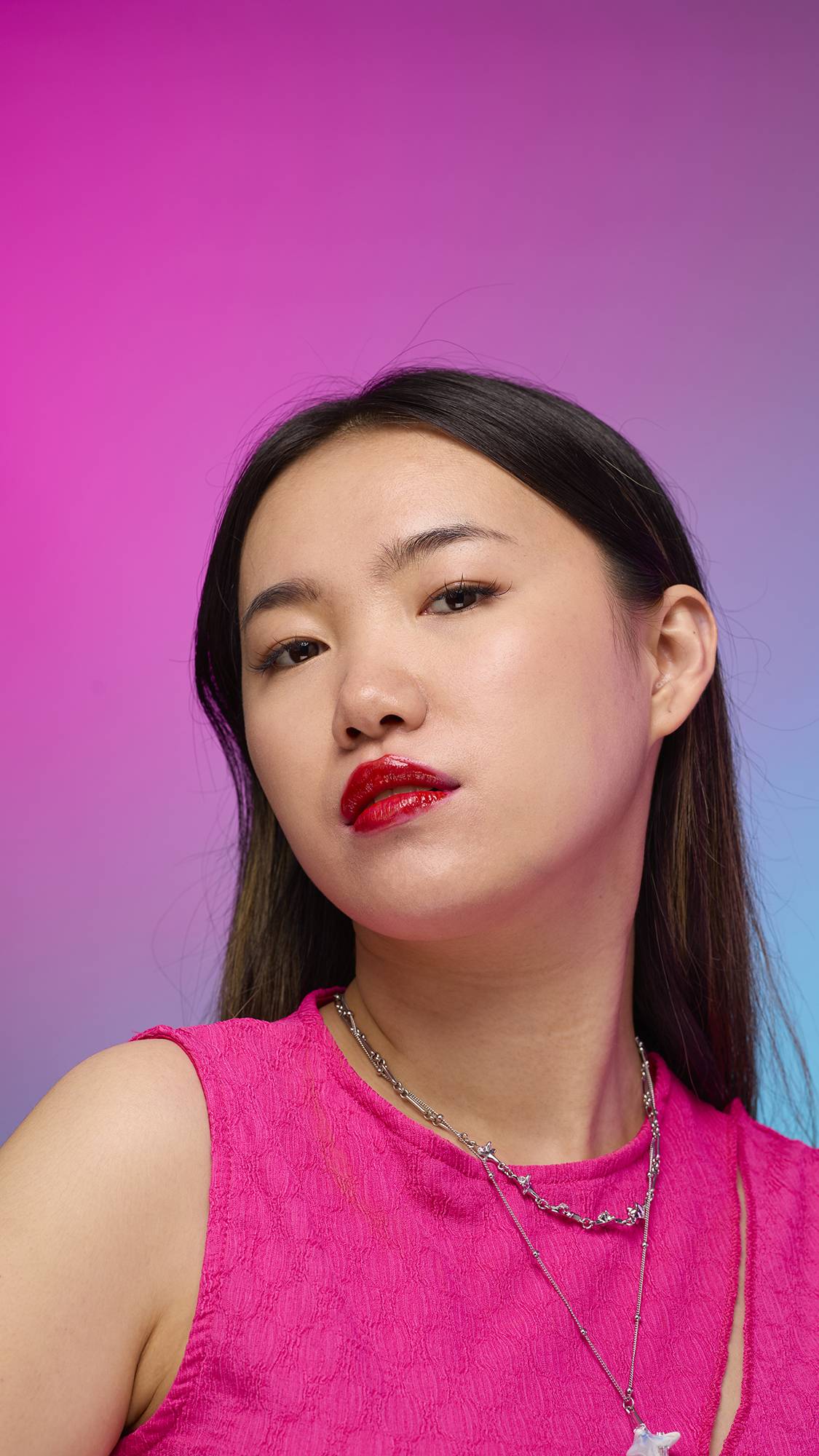 The model has long, straight dark hair and is on a pastel pink, blue and purple hue background. Their make-up has been done with the focal point being the juicy coating of cherry-red tint on their lips created by the Cherry lip jelly.