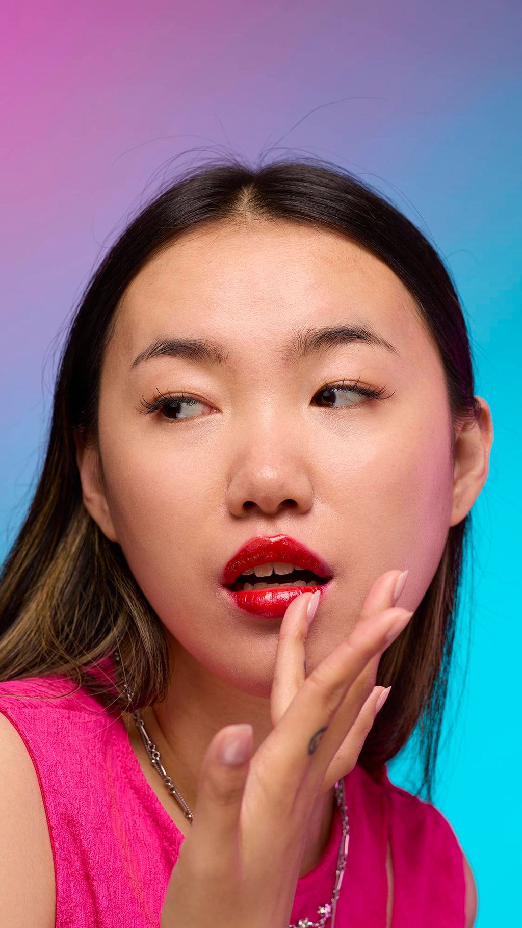 The model has long, dark hair and is on a pastel pink, blue and purple hue background. Their hand is by their face adjusting the juicy coating of blushed-red tint on their lips created by the Cherry lip jelly.