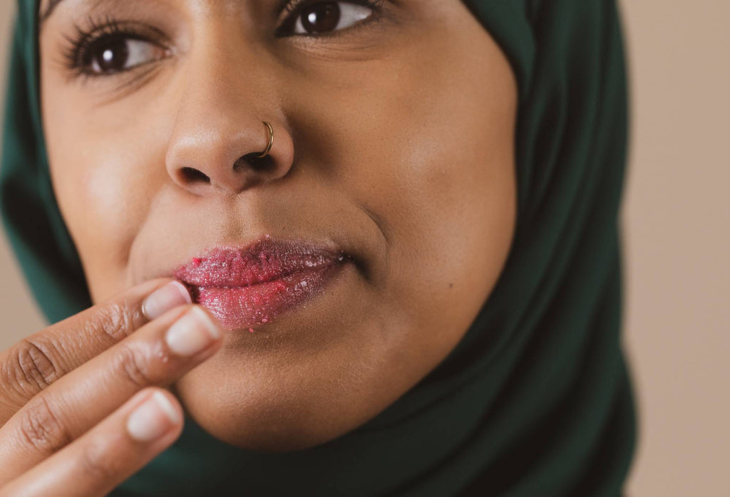 A person with a nose piercing, wearing a Hijab, scrubs their pouted lips with fuchsia pink Cherry sugar lip scrub.