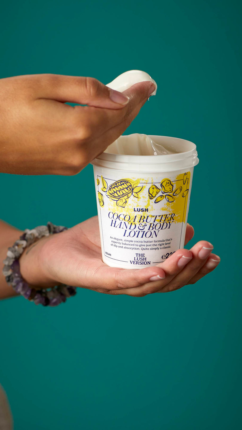 The image shows the model's arms stretched into the centre holding the white, LUSH Version-themed packed pot of the Cocoa Butter hand and body lotion. One of their hands is scooping the product out. There is a teal background.