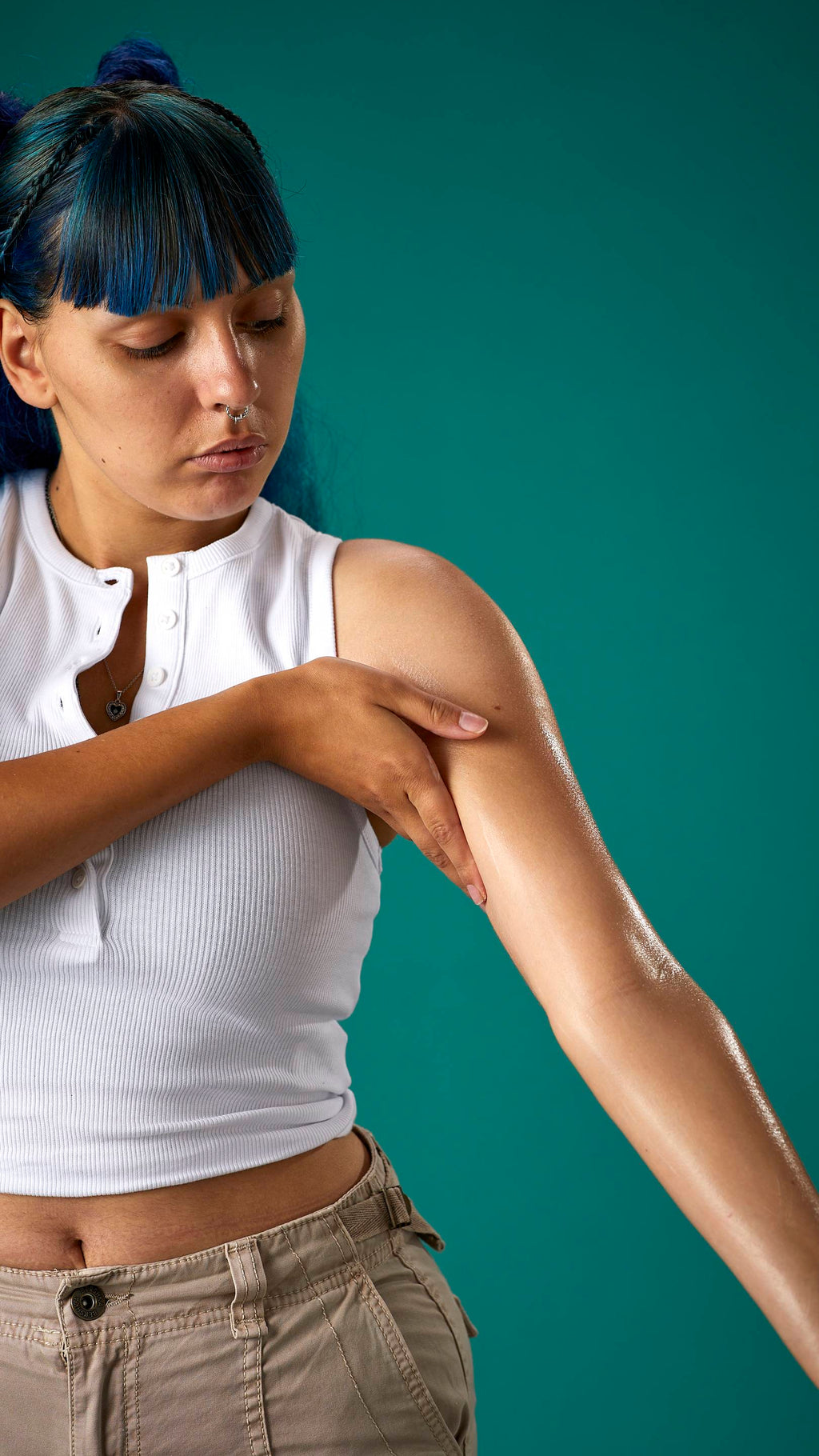 The image shows the model who has blue hair with a full fringe and space buns. They are wearing a white cropped top and high-waisted trousers, on a teal background. They are gently soothing the Cocoa Butter lotion to their arm.