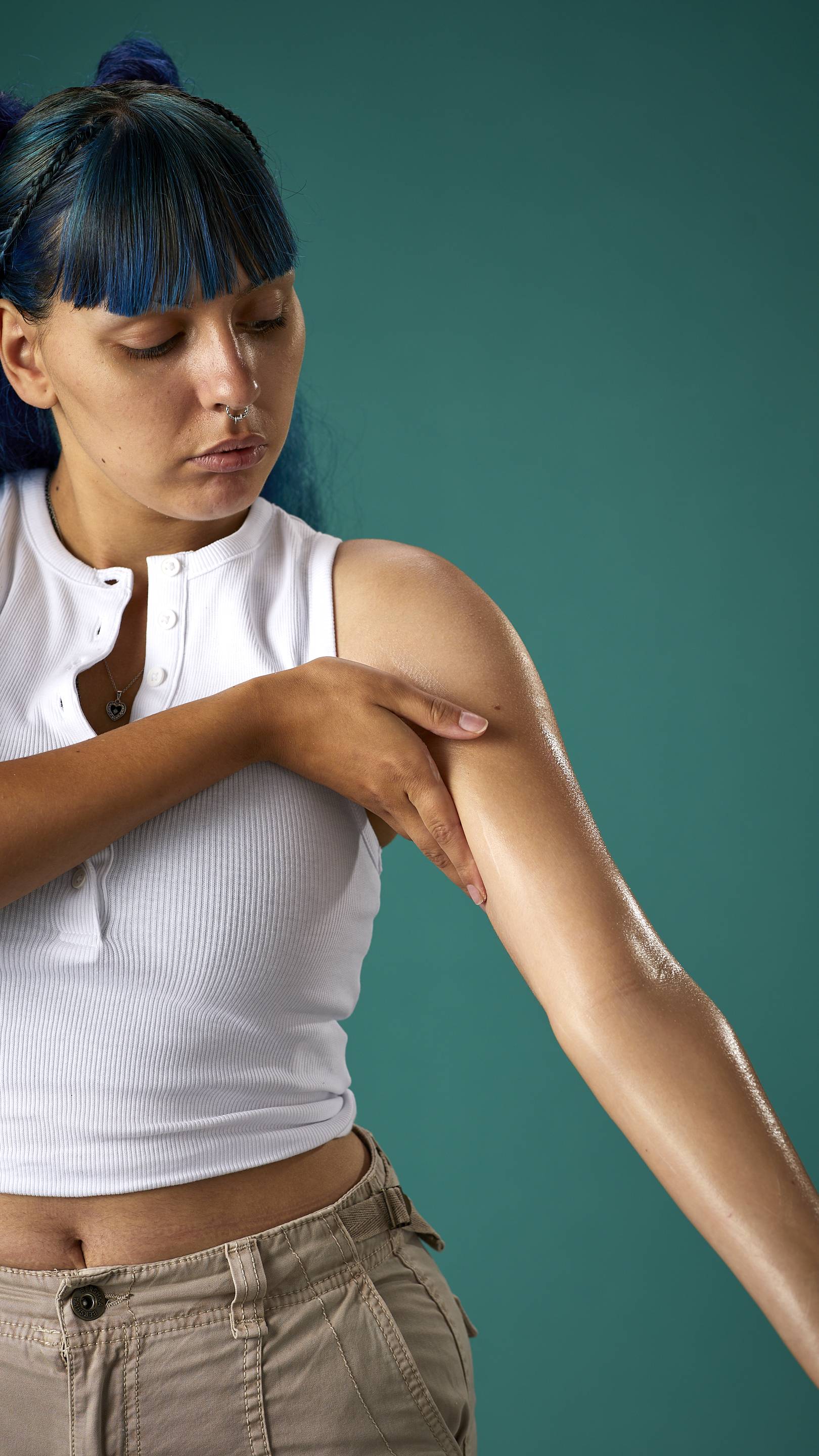 The image shows the model who has blue hair with a full fringe and space buns. They are wearing a white cropped top and high-waisted trousers, on a teal background. They are gently soothing the Cocoa Butter lotion to their arm.