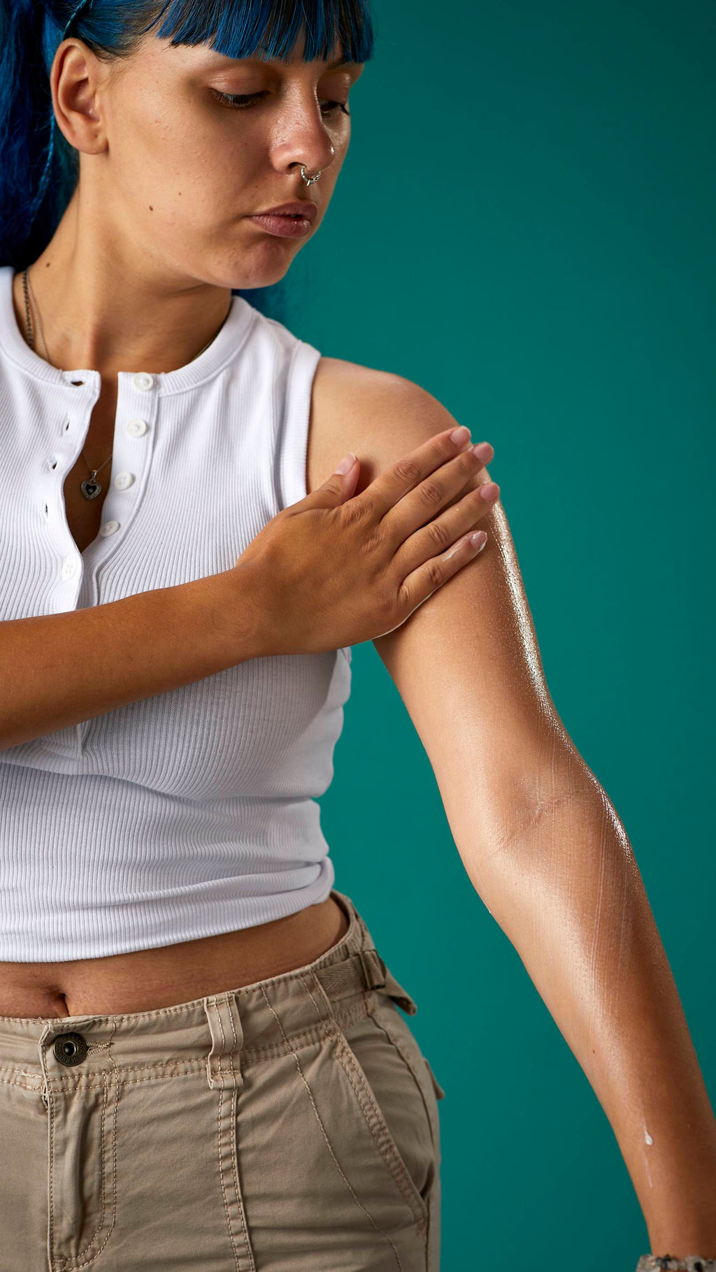 The image shows a close-up of half the model's torso. They are wearing a white cropped top and high-waisted trousers, on a teal background. They are gently soothing the Cocoa Butter lotion to their arm.