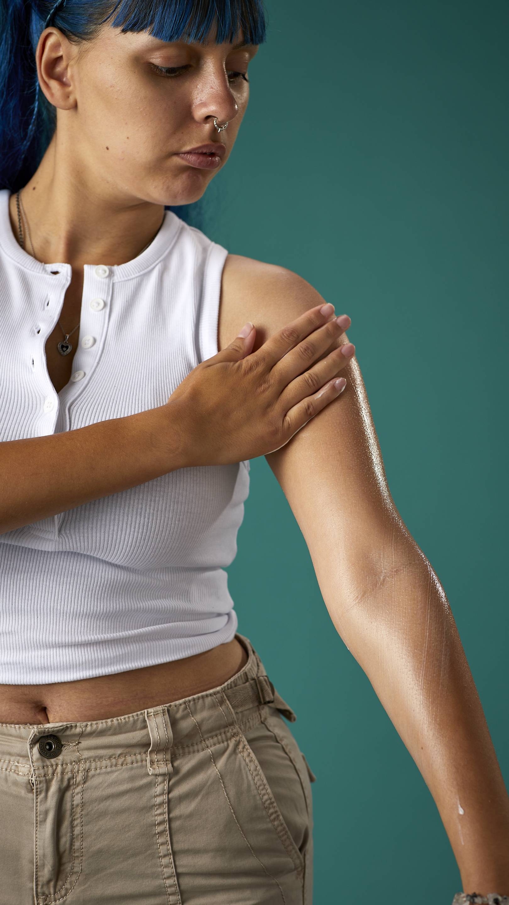 The image shows a close-up of half the model's torso. They are wearing a white cropped top and high-waisted trousers, on a teal background. They are gently soothing the Cocoa Butter lotion to their arm.
