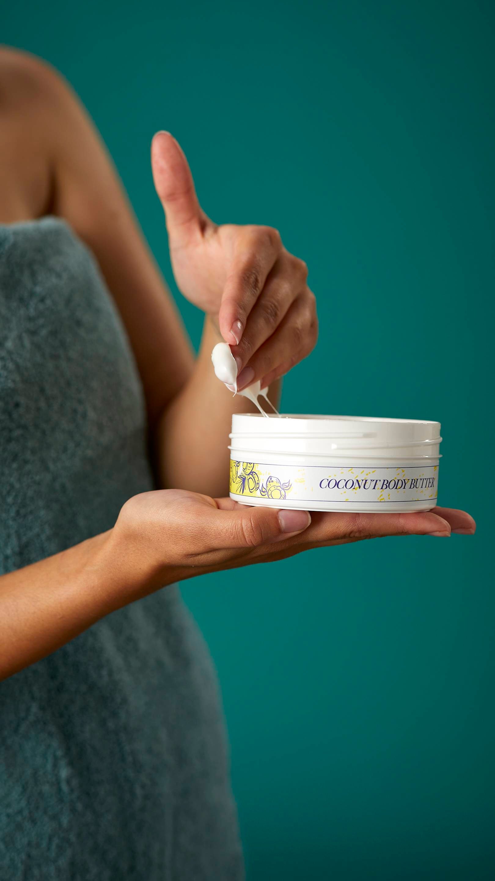 A close-up shot of the model's hands. One hand is holding the flat round, white, LUSH Version-themed packed tub of Coconut body butter.
