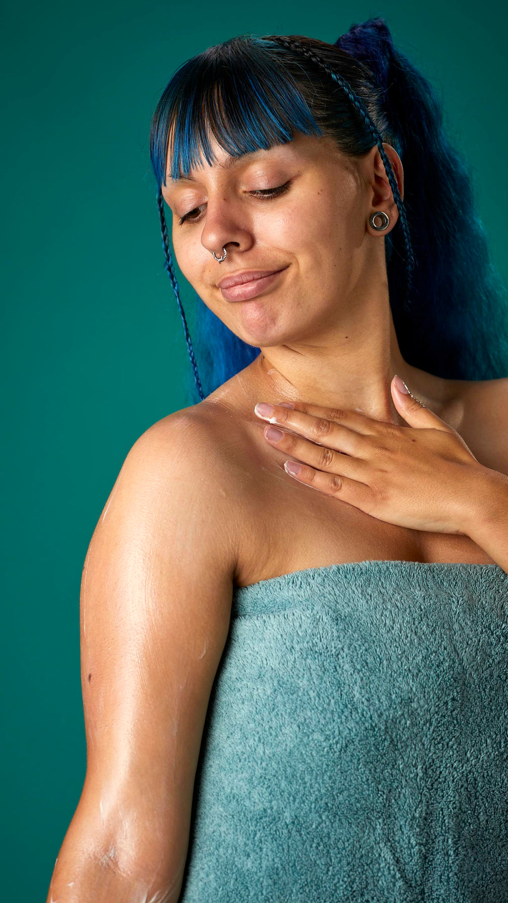 The image shows the model wrapped in a sage-green towel on a teal background. They have blue hair with a full fringe and are gently applying the Coconut body butter to their upper arm.