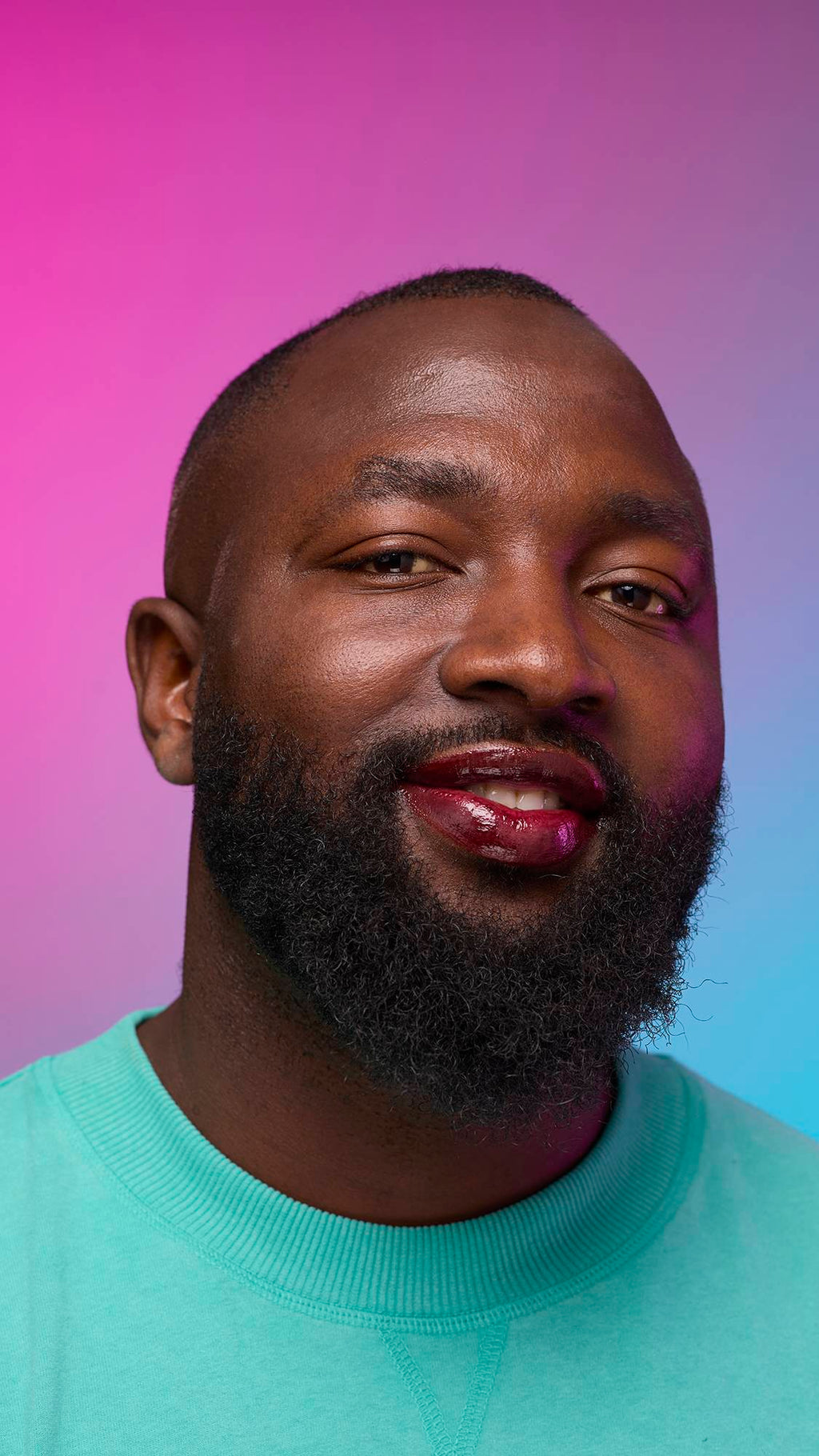 The model is on a pastel pink, blue and purple hue background in a vivid teal jumper. They are glancing away with the focal point being the juicy coating of grounding, earth-toned tint on their lips created by the Cola lip jelly.