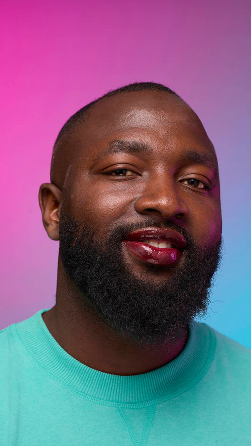 The model is on a pastel pink, blue and purple hue background in a vivid teal jumper. They are glancing away with the focal point being the juicy coating of grounding, earth-toned tint on their lips created by the Cola lip jelly.