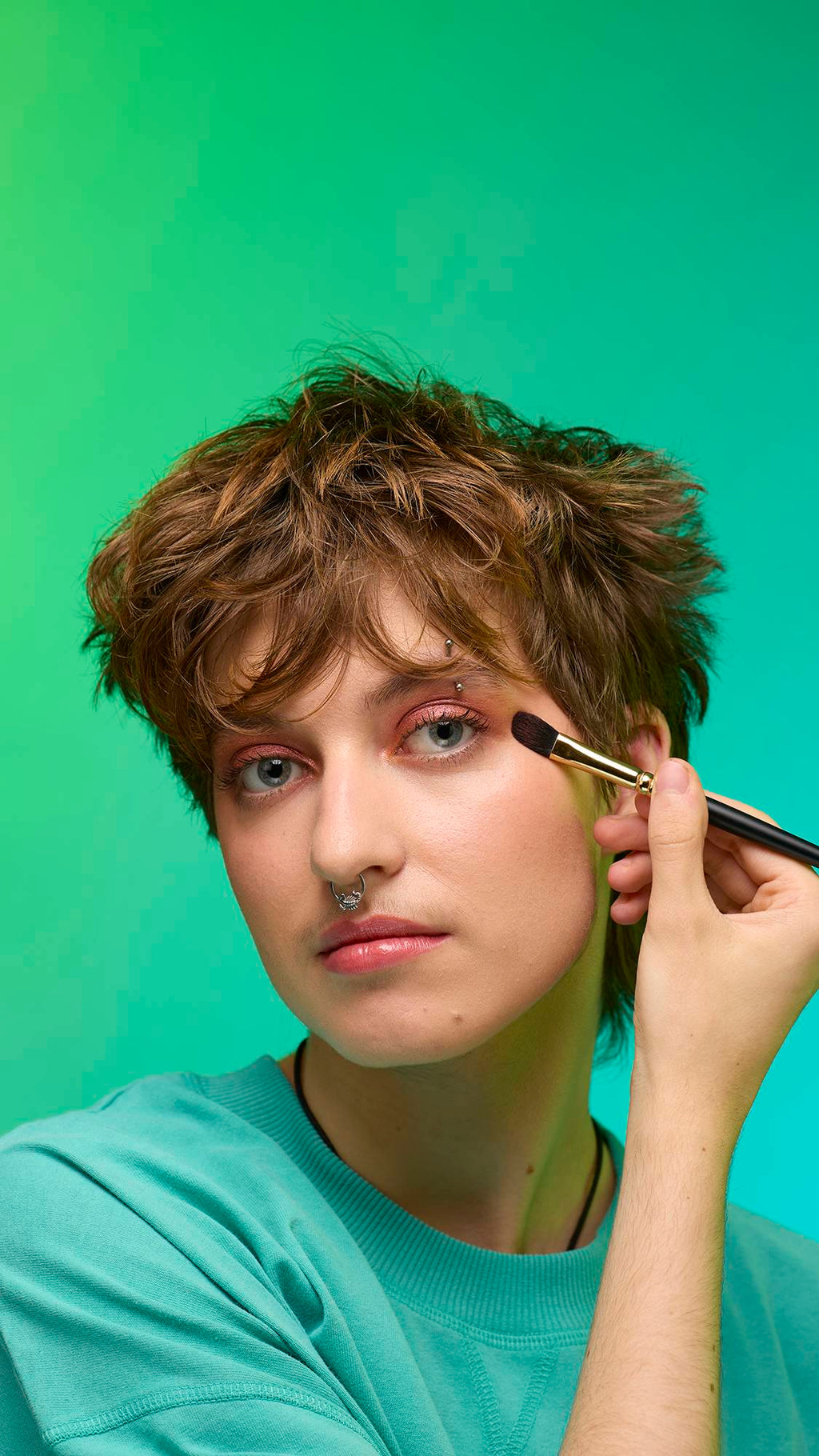 The image shows a close-up of the model's face on a pale blue background. They are holding a make-up brush to their eye area as they apply the shimmering, bronze, Glitter Crumble to their eyelids.