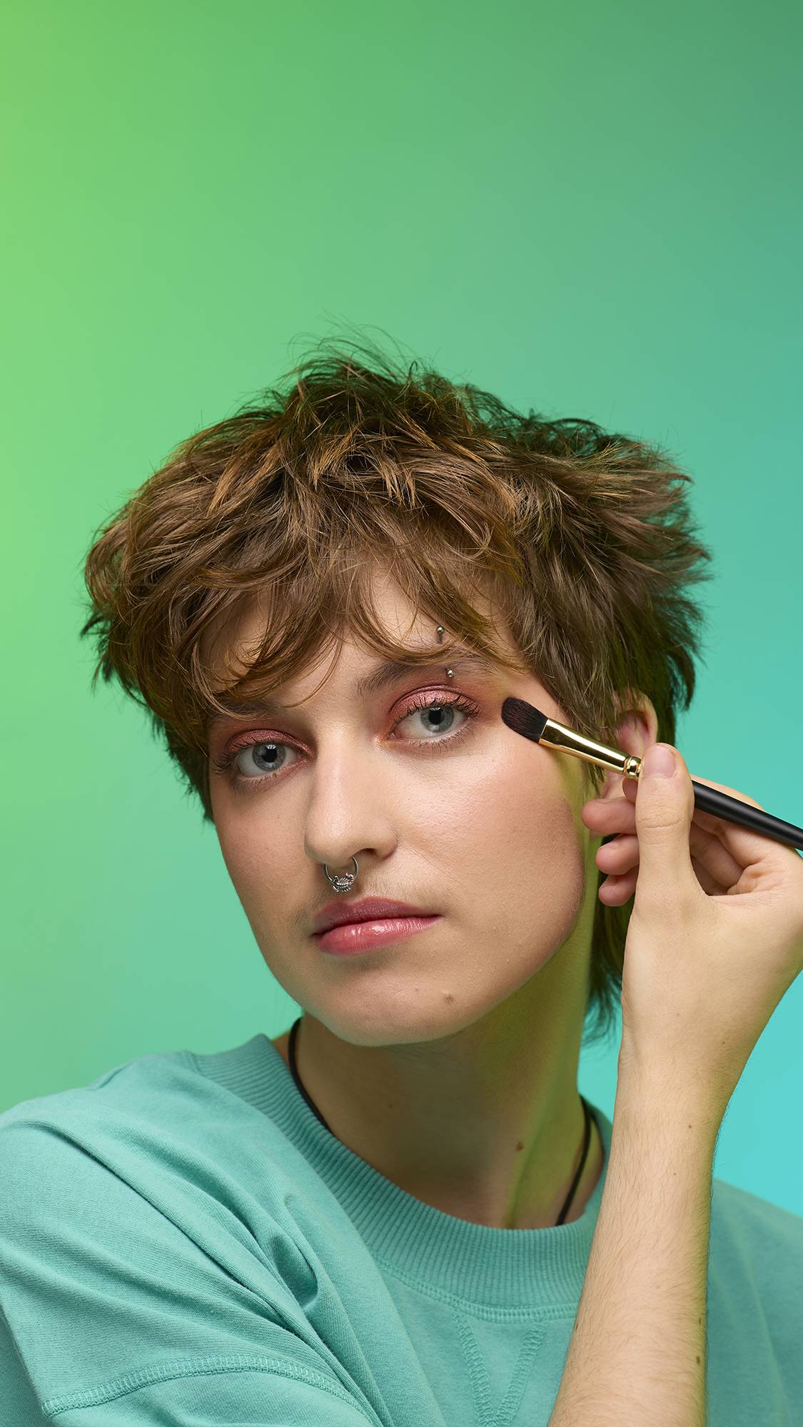 The image shows a close-up of the model's face on a pale blue background. They are holding a make-up brush to their eye area as they apply the shimmering, bronze, Glitter Crumble to their eyelids.
