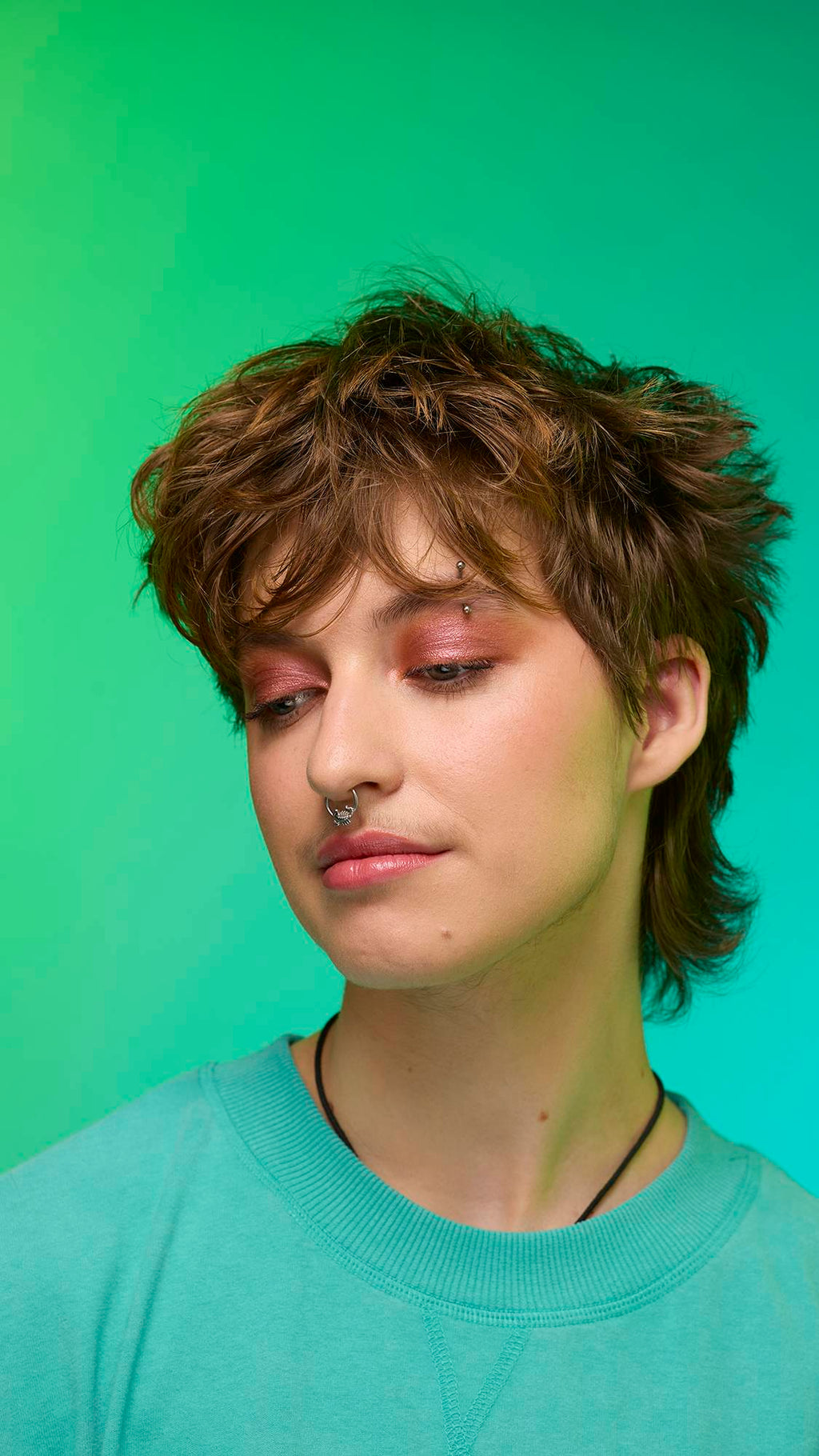 The image shows a close-up of the model's face on a pale blue-to-green gradient background. The model's make-up is done with the focal point being a shimmering, metallic-bronze eyeshadow made by the Comet Glitter Crumble.