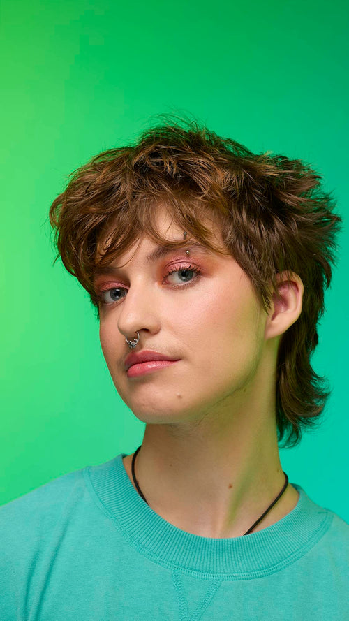 The image shows a close-up of the model's face on a pale blue-to-green gradient background. The model's make-up is done with the focal point being a shimmering, metallic-bronze eyeshadow made by the Comet Glitter Crumble.