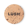 Cork Pot. A round, beige, product holder made of natural cork. LUSH is printed on top, in black, bold letters.