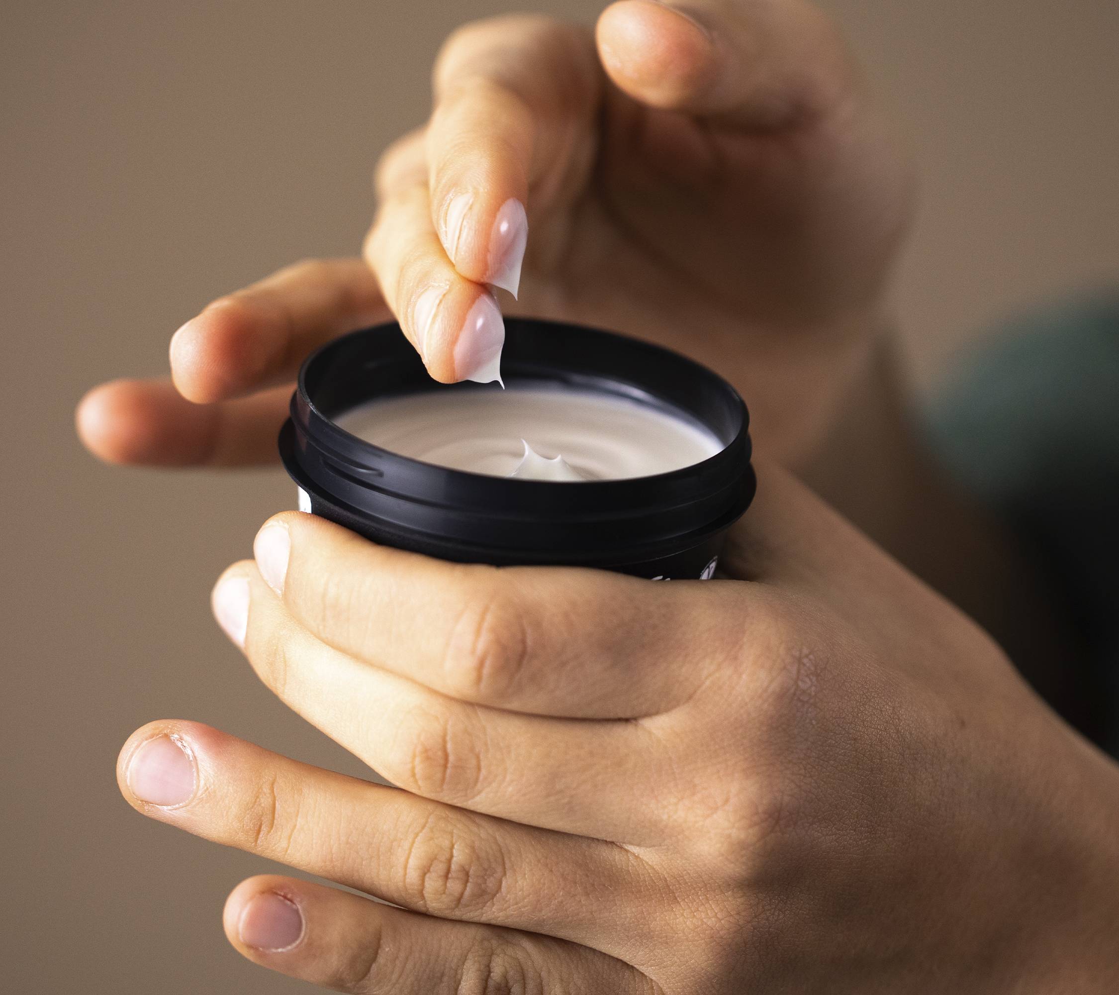 A close-up of hands holding the Cosmetic Lad pot with one hand dipping two fingers into the velvety, creamy moisturiser.