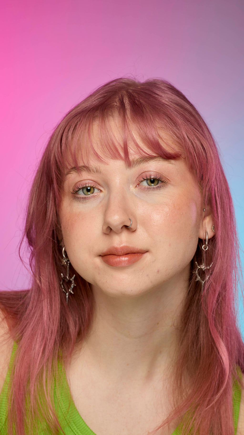 The image shows a fair-skinned model on a pink and blue gradient background. They have pink hair and subtle shimmering lips from using the Crackled Copper lip cream.