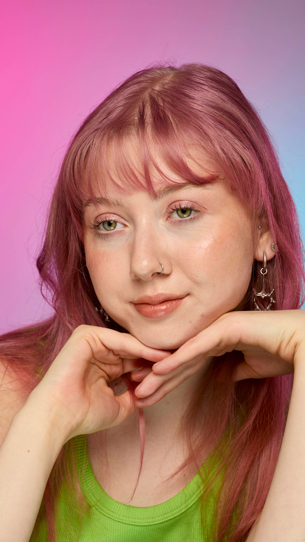 The image shows a fair-skinned model on a pink and blue gradient background as they rest their chin on their hands. They have pink hair and subtle shimmering lips from using the Crackled Copper lip cream.