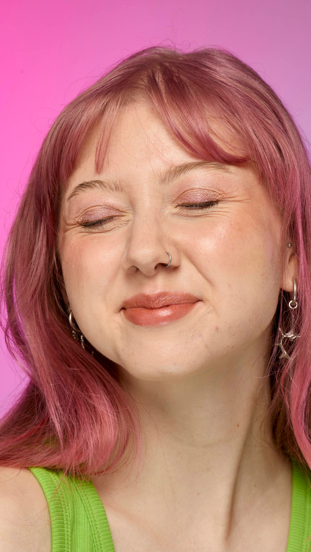 The image shows a fair-skinned model on a pink and blue gradient background, smiling with their eyes closed. They have pink hair and subtle shimmering lips from using the Crackled Copper lip cream.