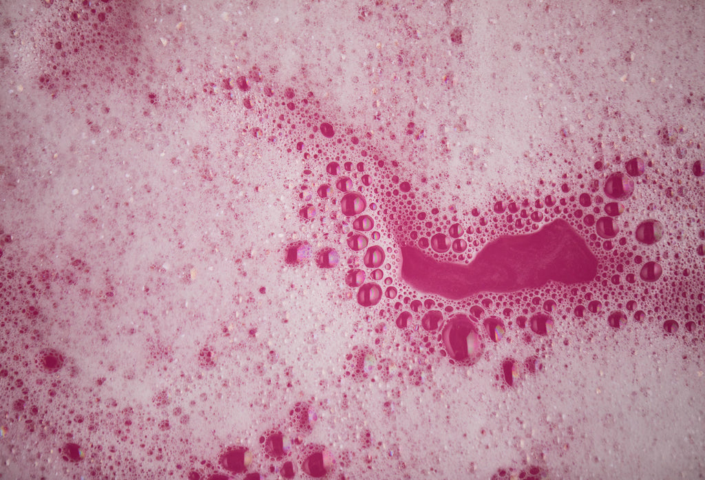 Image shows soft, foamy bubbles on a deep pink sea of bath water.